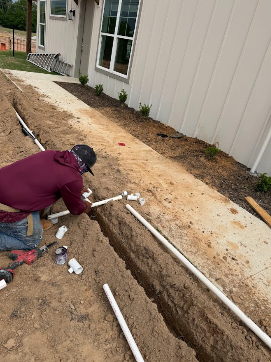Worker installing PVC pipes in a trench beside a house with beige siding and a concrete walkway.