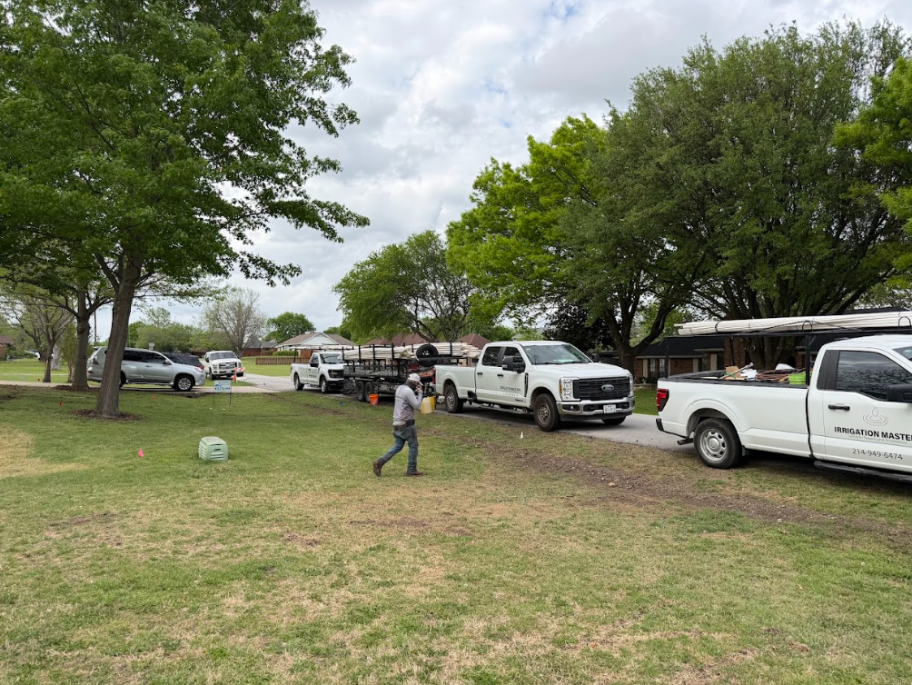 Worker walking on grass near trucks parked along a tree-lined street under a cloudy sky.