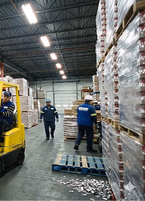 Warehouse workers in safety helmets and uniforms managing pallets of wrapped goods with a forklift in a spacious industrial storage area.