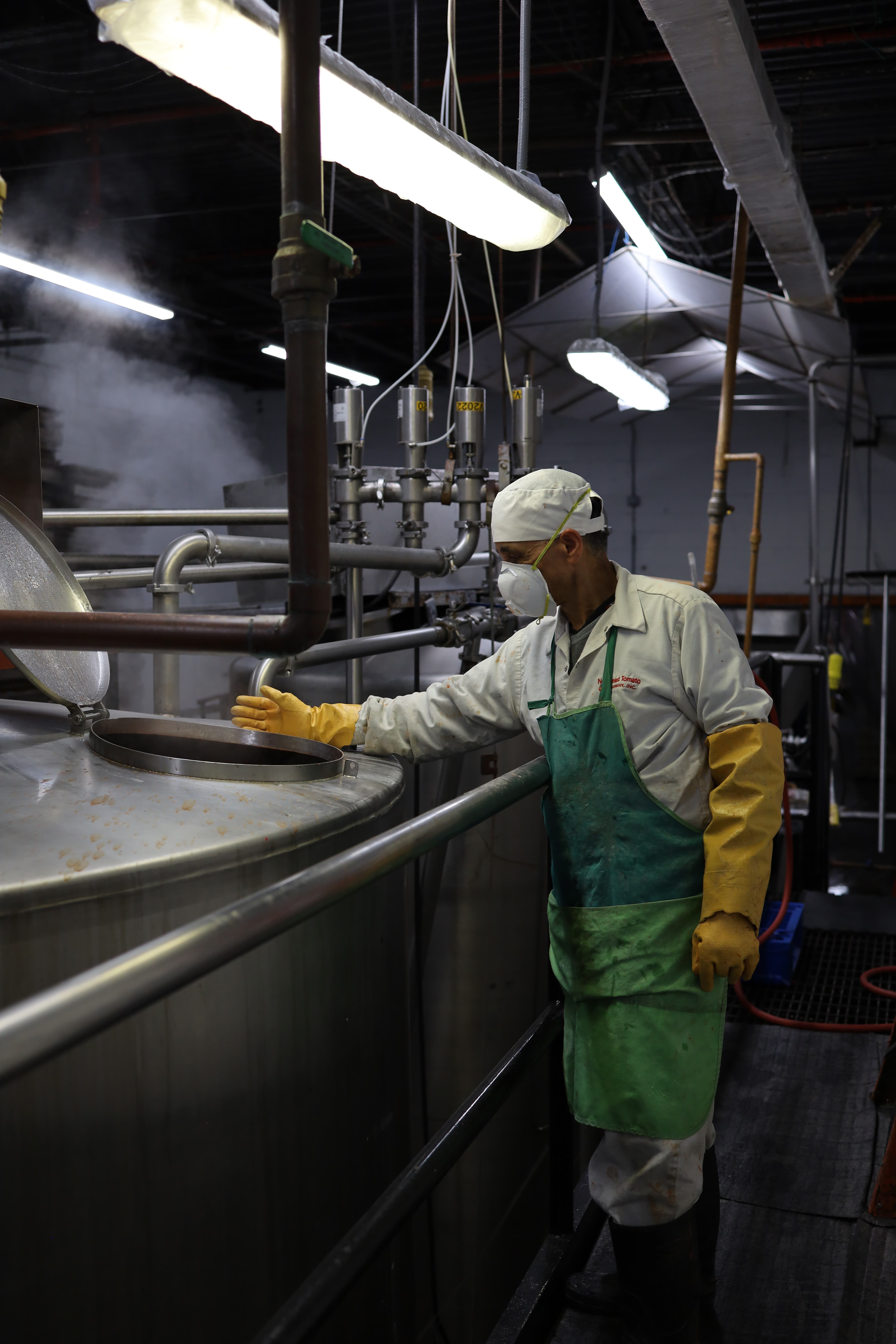 Worker wearing protective gloves, mask, and apron inspecting steam rising from a large industrial stainless steel tank in a factory.