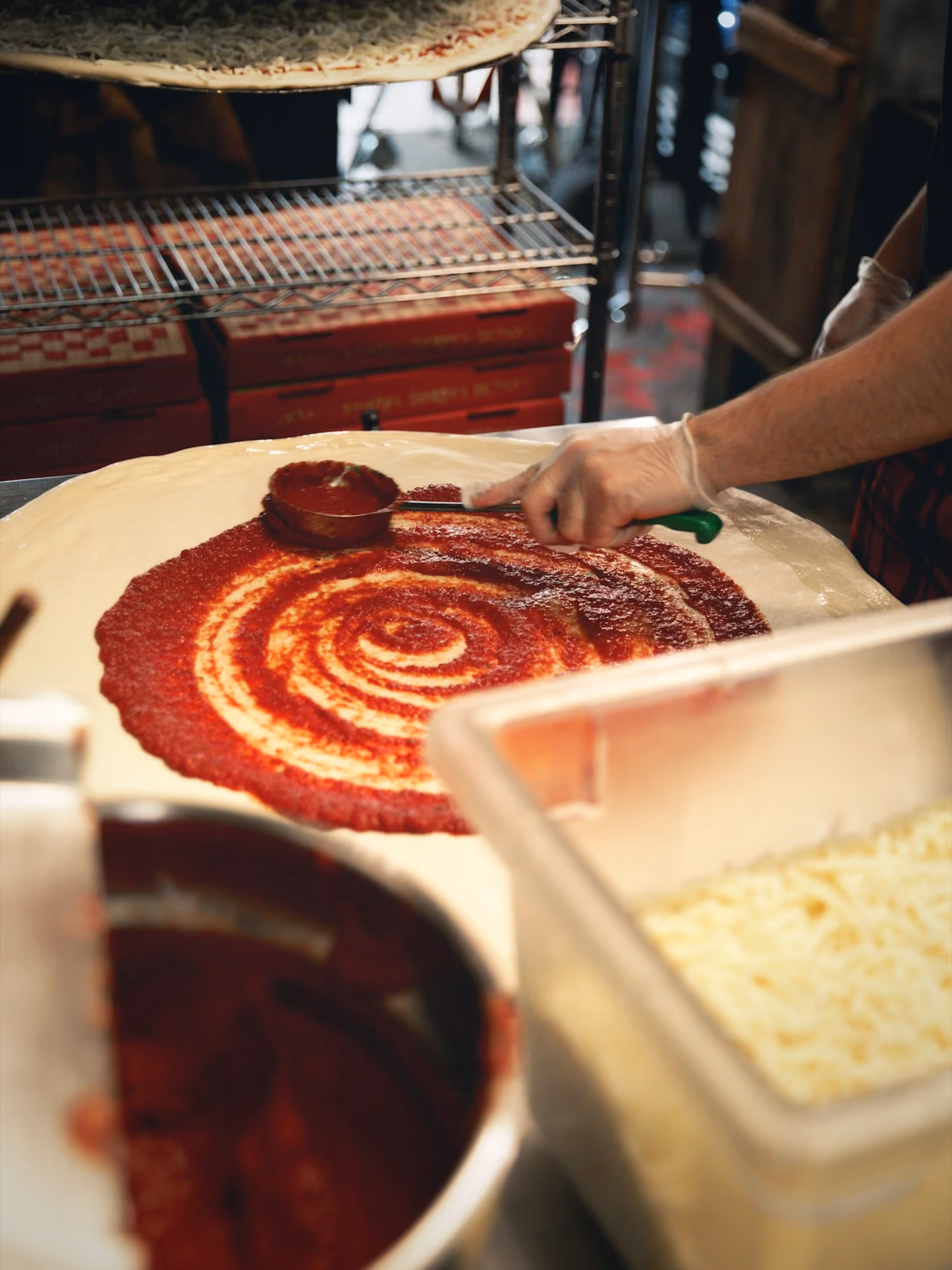 Person spreading tomato sauce on large pizza dough in a kitchen setting.