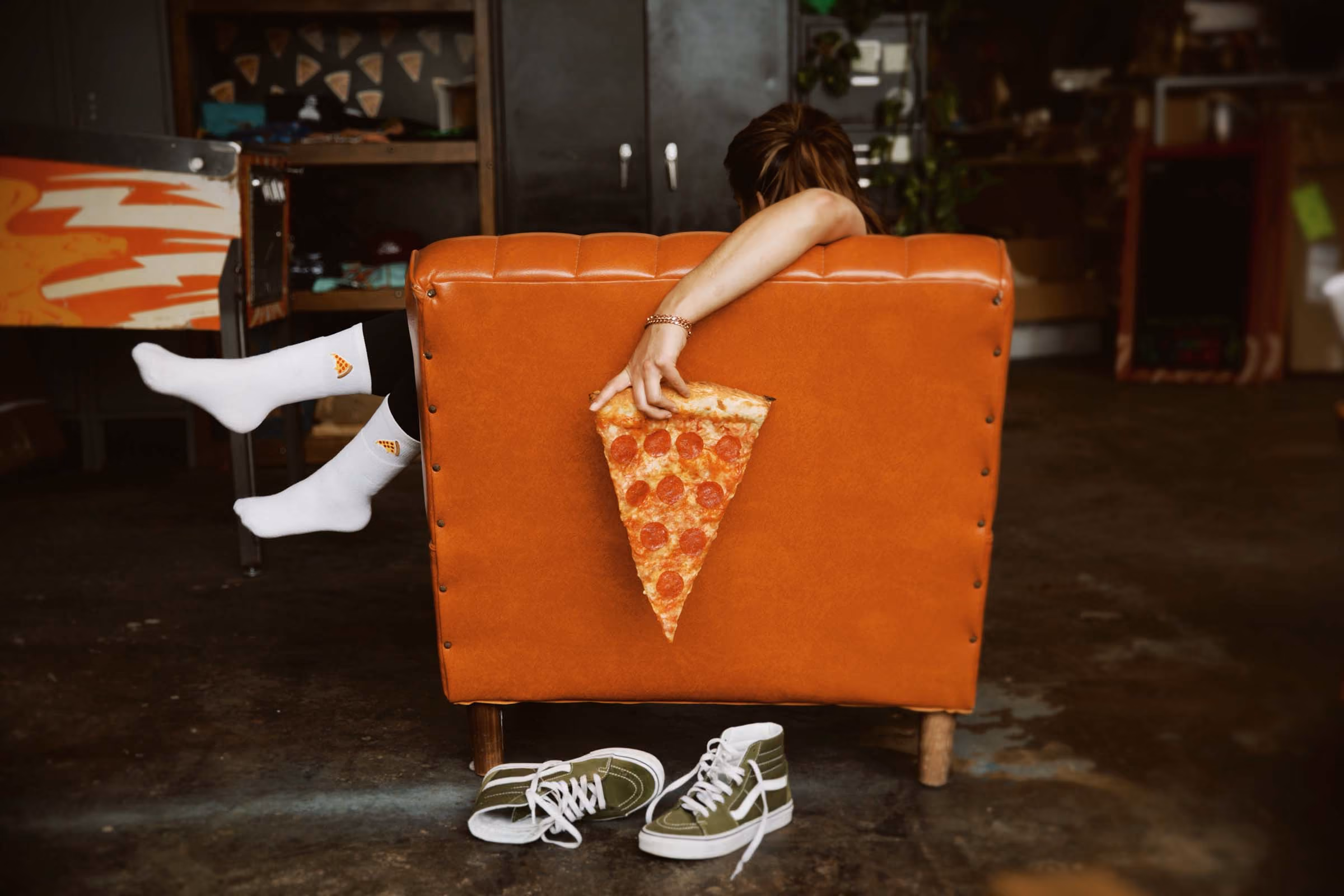 Person in white pizza socks sits on orange chair holding large pepperoni pizza slice behind back.