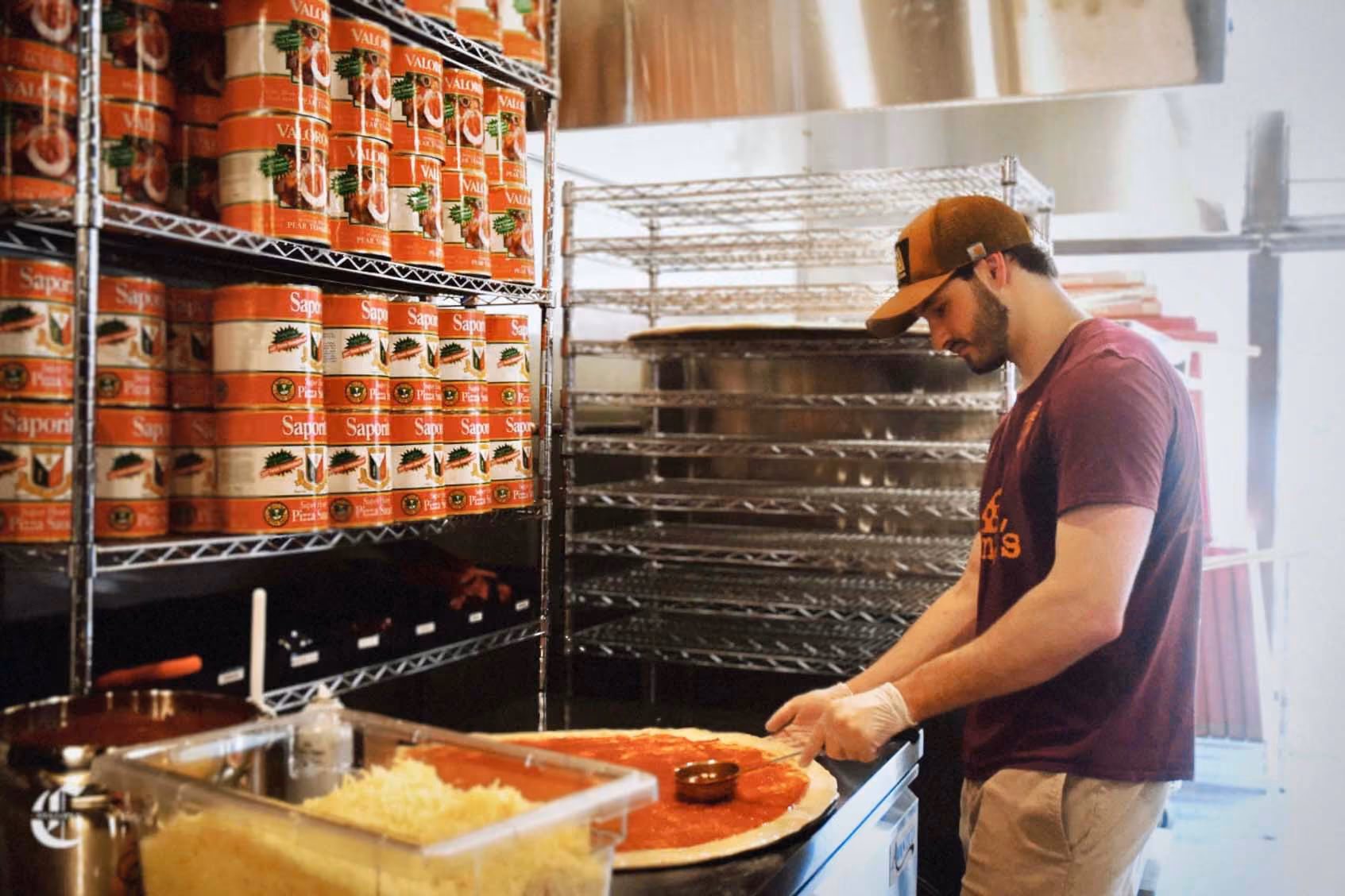 Man in a cap spreading tomato sauce on a large pizza dough in a kitchen with canned tomatoes on shelves.