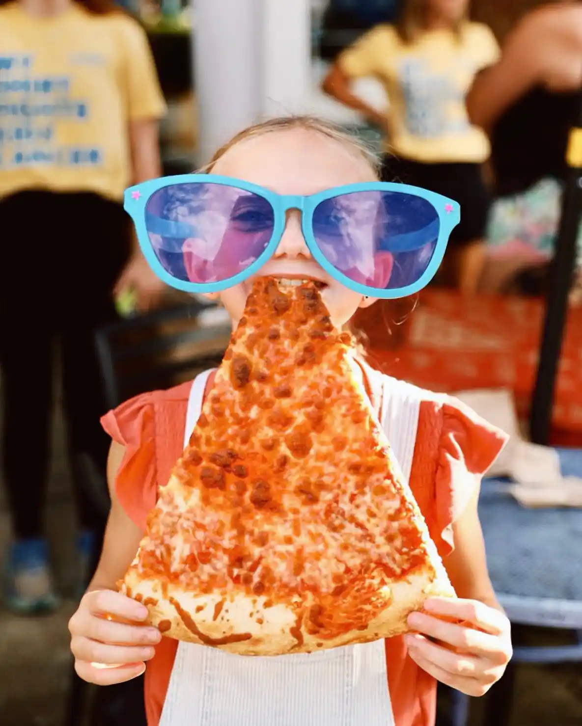 Child wearing oversized blue sunglasses holds and bites a large slice of cheese pizza.