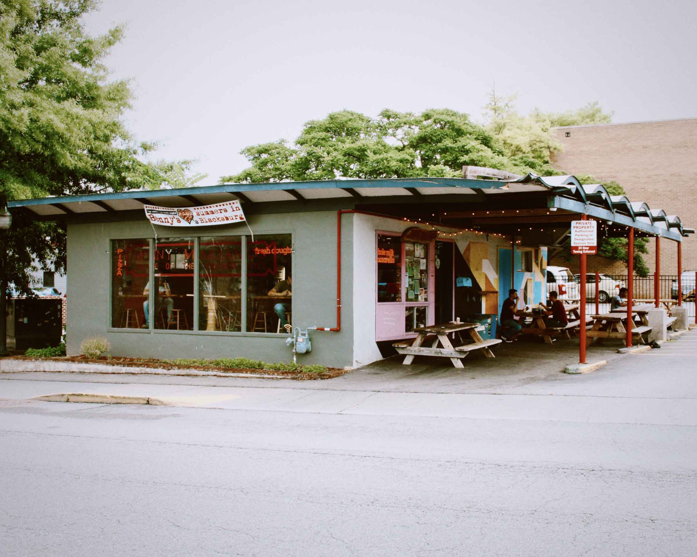 Small pizzeria with outdoor picnic tables under a roof and people dining outside.