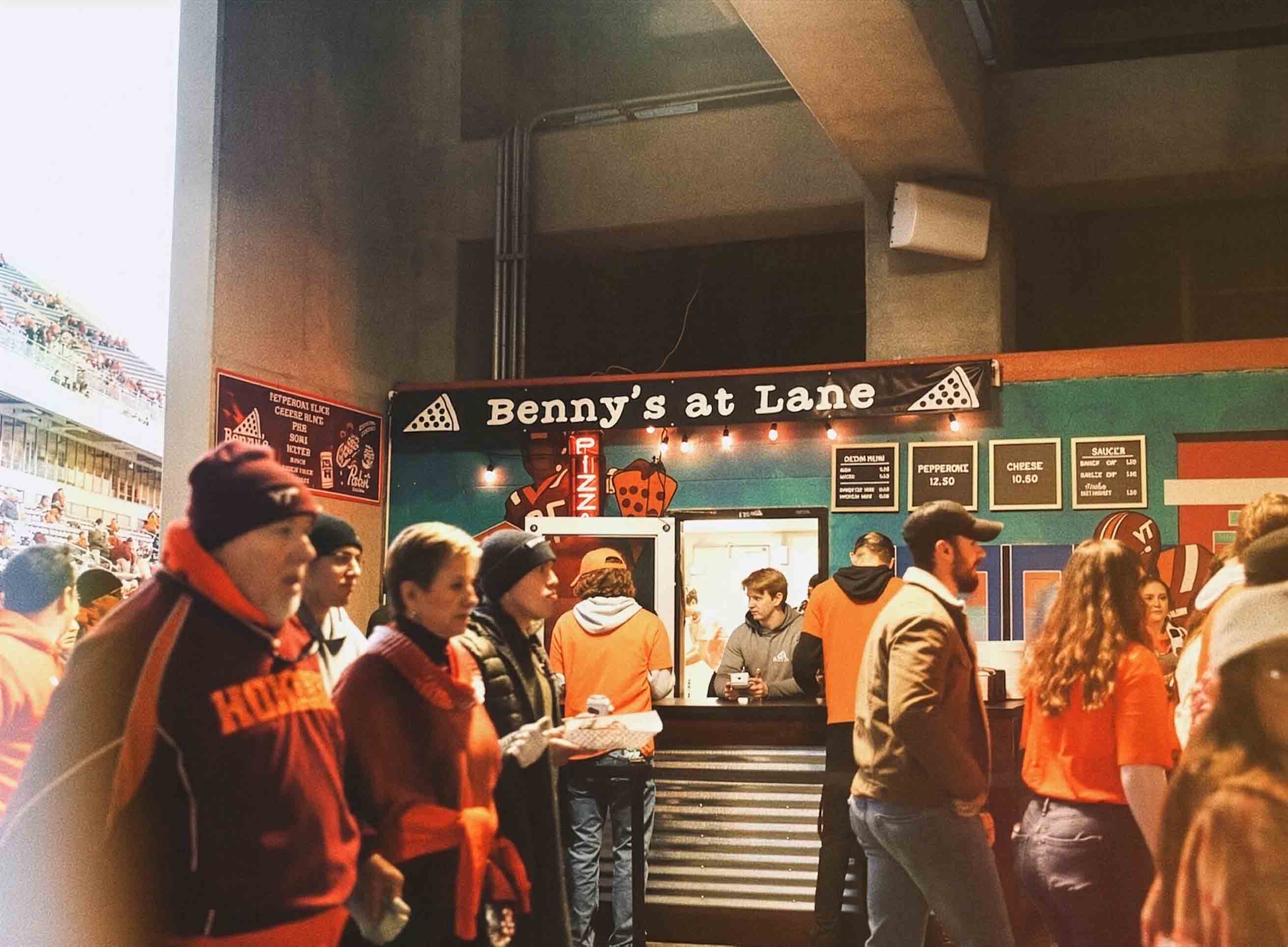 People lined up at Benny's at Lane pizza stand inside a stadium with menu signs overhead.