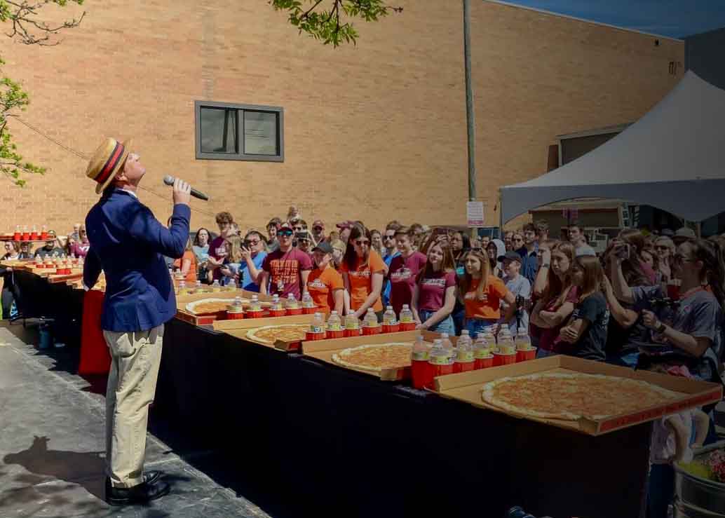 Man in a hat and blue blazer speaking into a microphone to an audience near large pizzas outdoors.