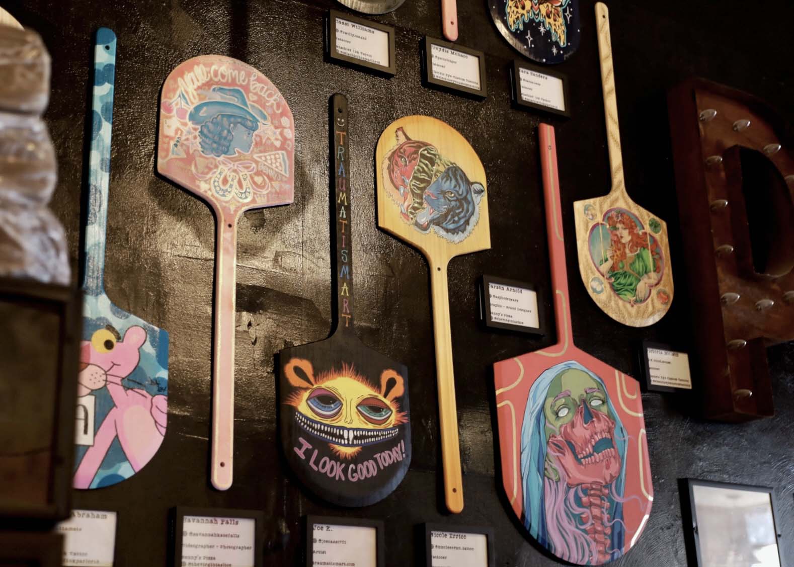 Wall display of pizza paddles with colorful, artistic painted faces and designs.