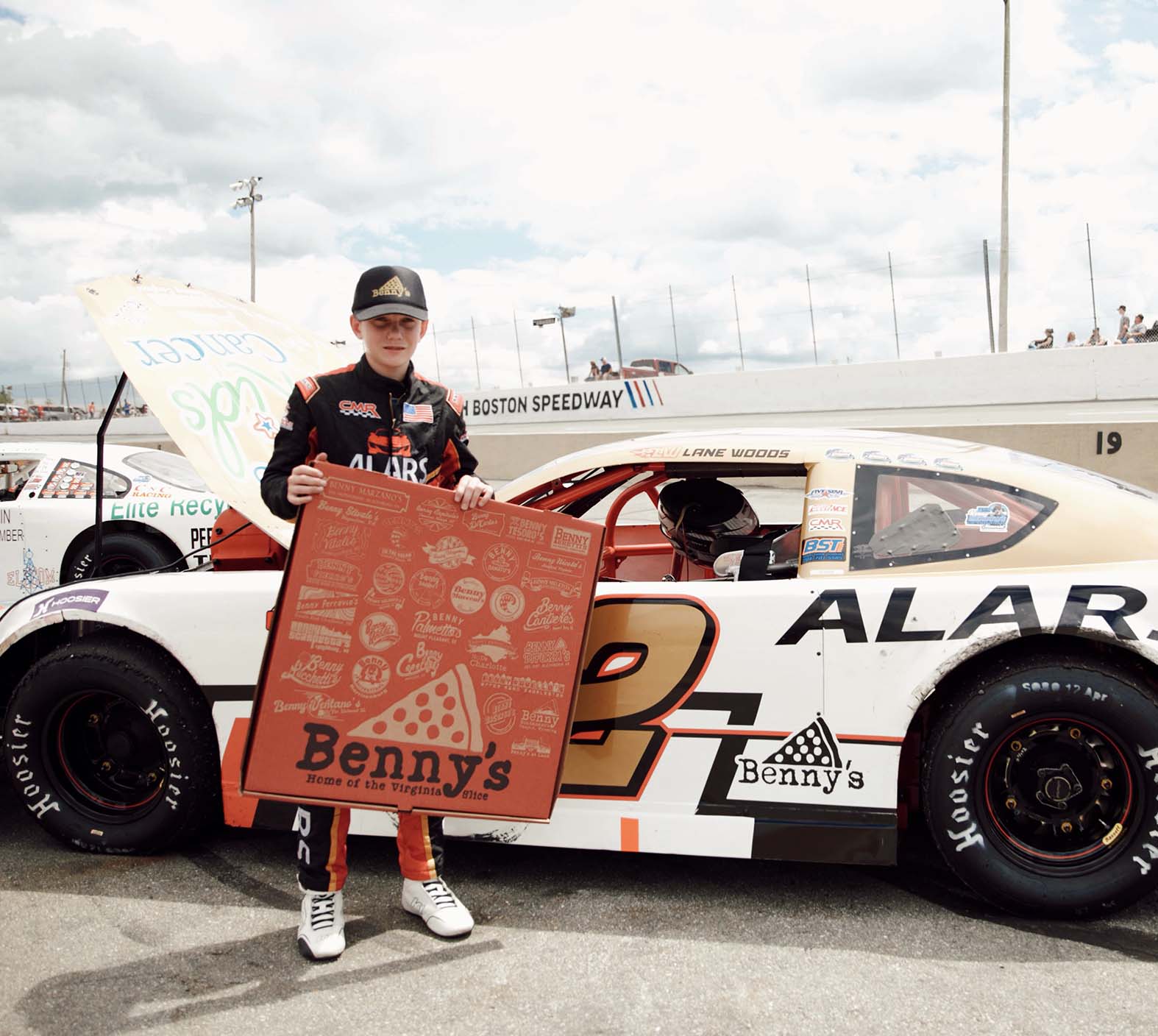 Young race car driver in suit holds a large Benny's pizza box beside car number 2 at Boston Speedway.