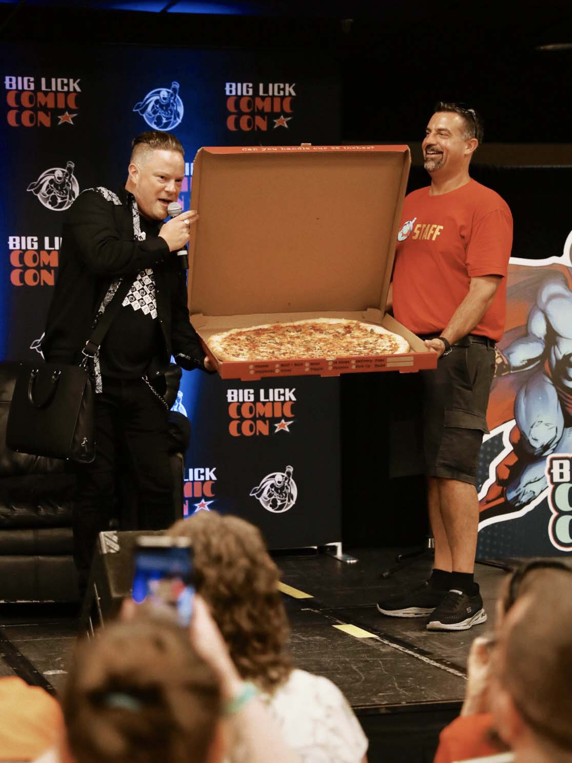 Two men on stage at Big Lick Comic Con holding and presenting a large pizza in an open box.