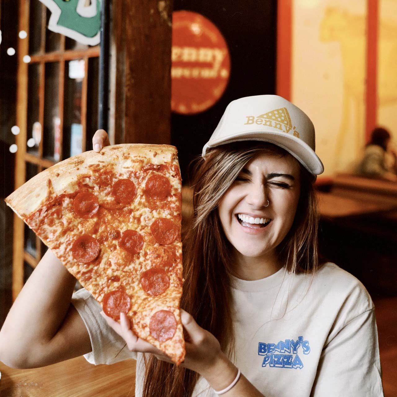 Woman in Benny's Pizza shirt and hat smiling while holding a large slice of pepperoni pizza.