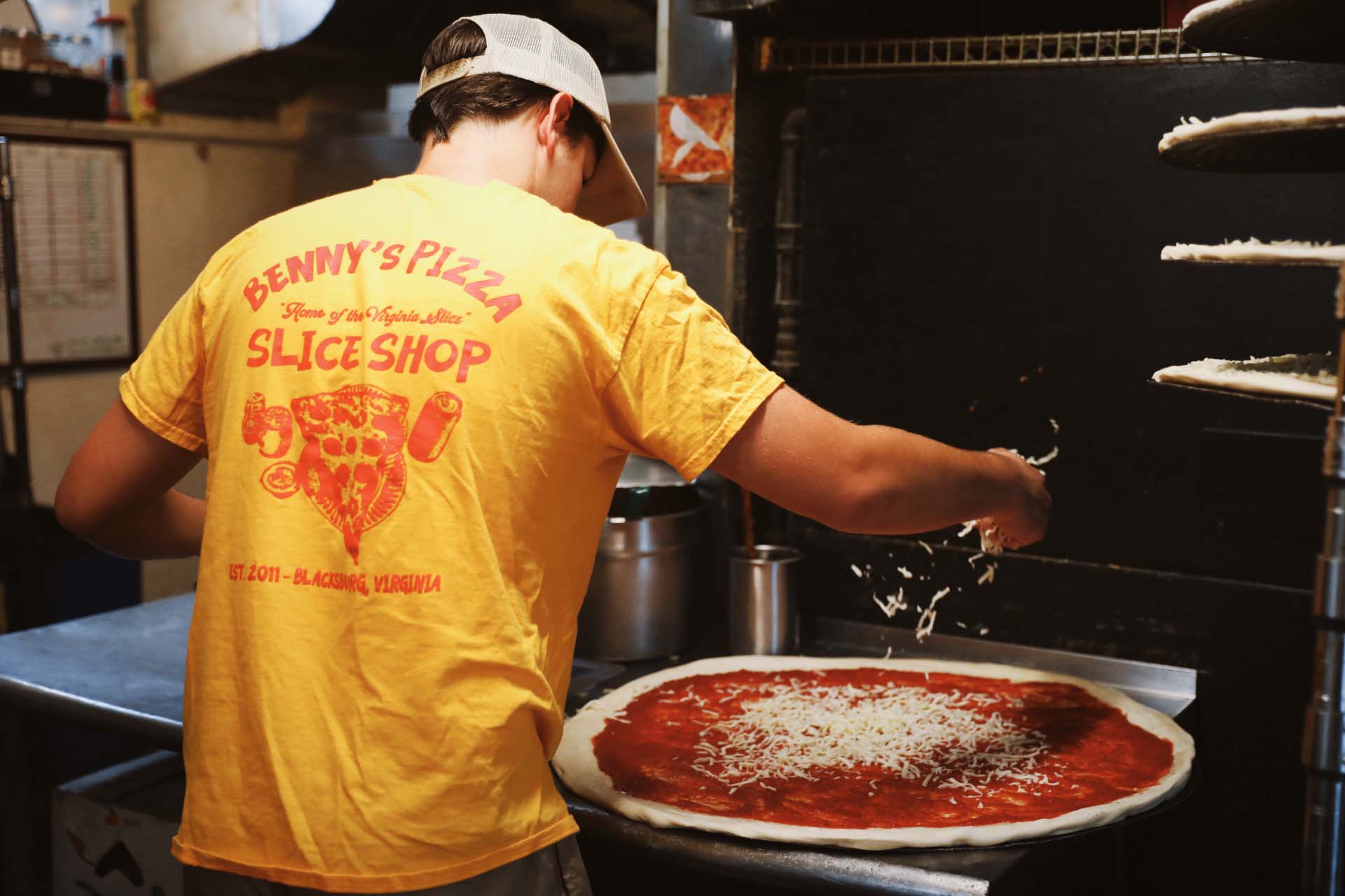 Person wearing yellow Benny's Pizza shirt sprinkling cheese on large pizza with tomato sauce.
