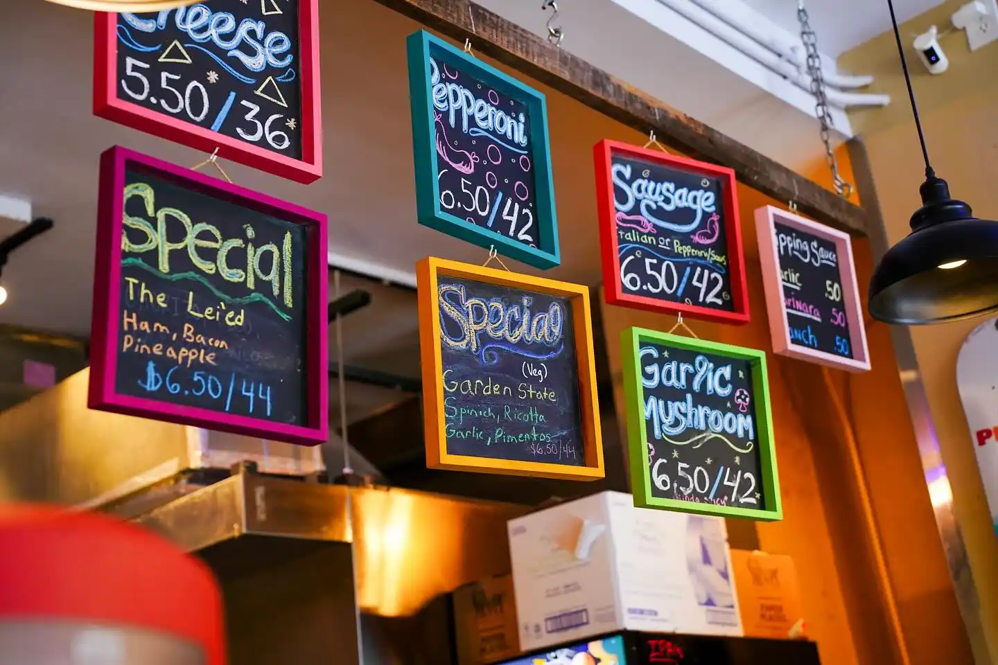 Colorful chalkboard menu signs hanging, listing pizza specials and prices in a restaurant kitchen.