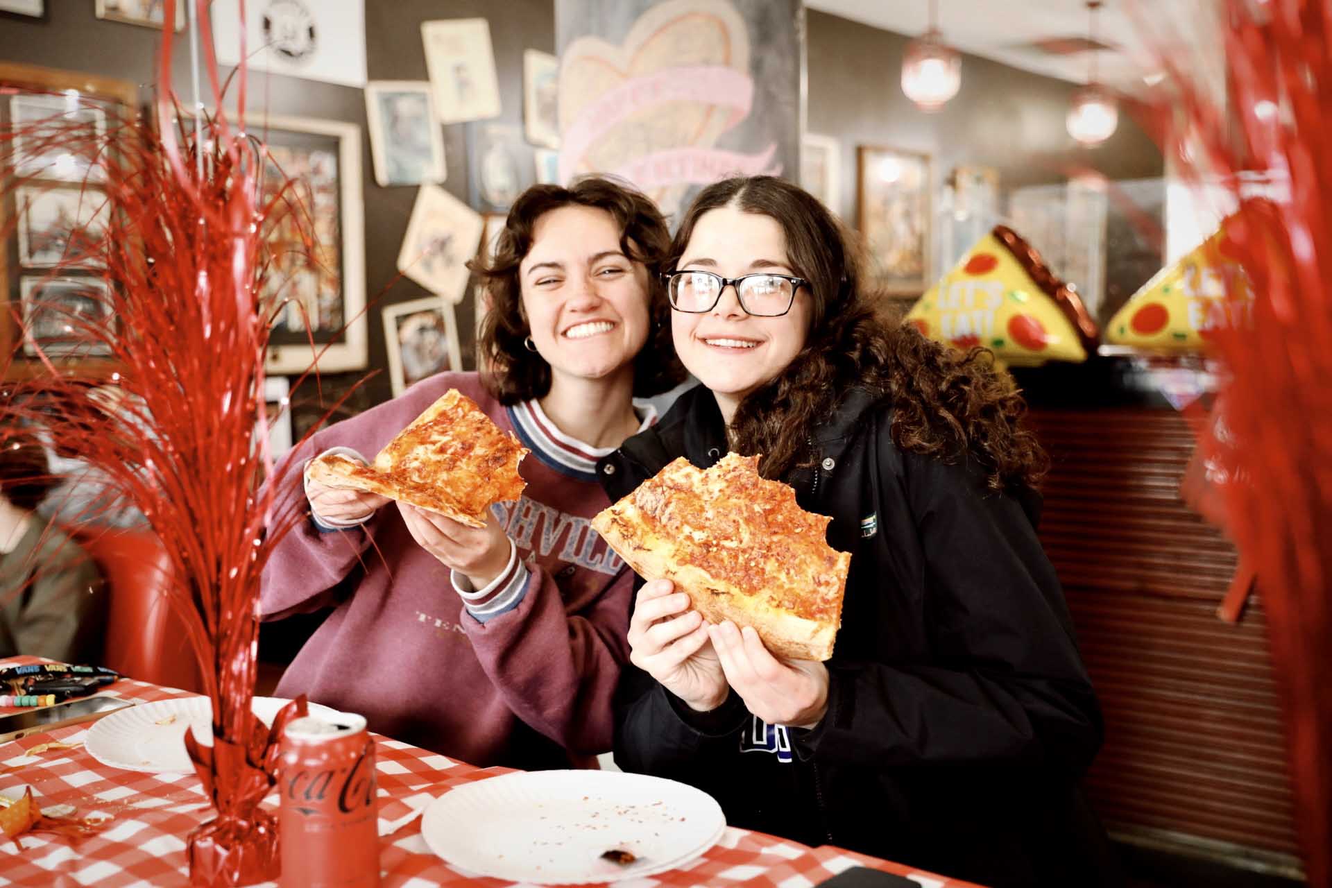 Two smiling women holding large slices of pizza in a cozy restaurant with red checkered tablecloth.