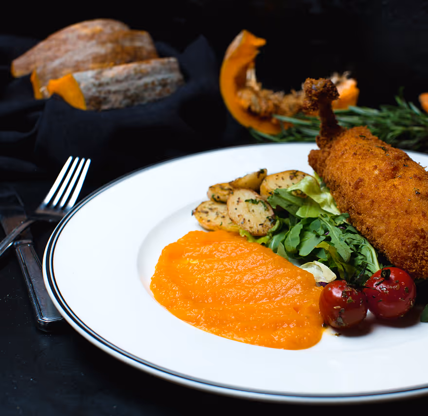 Plate with breaded fried chicken leg, mashed carrot, roasted potatoes, mixed greens, and cherry tomatoes, with sliced pumpkin in the background.