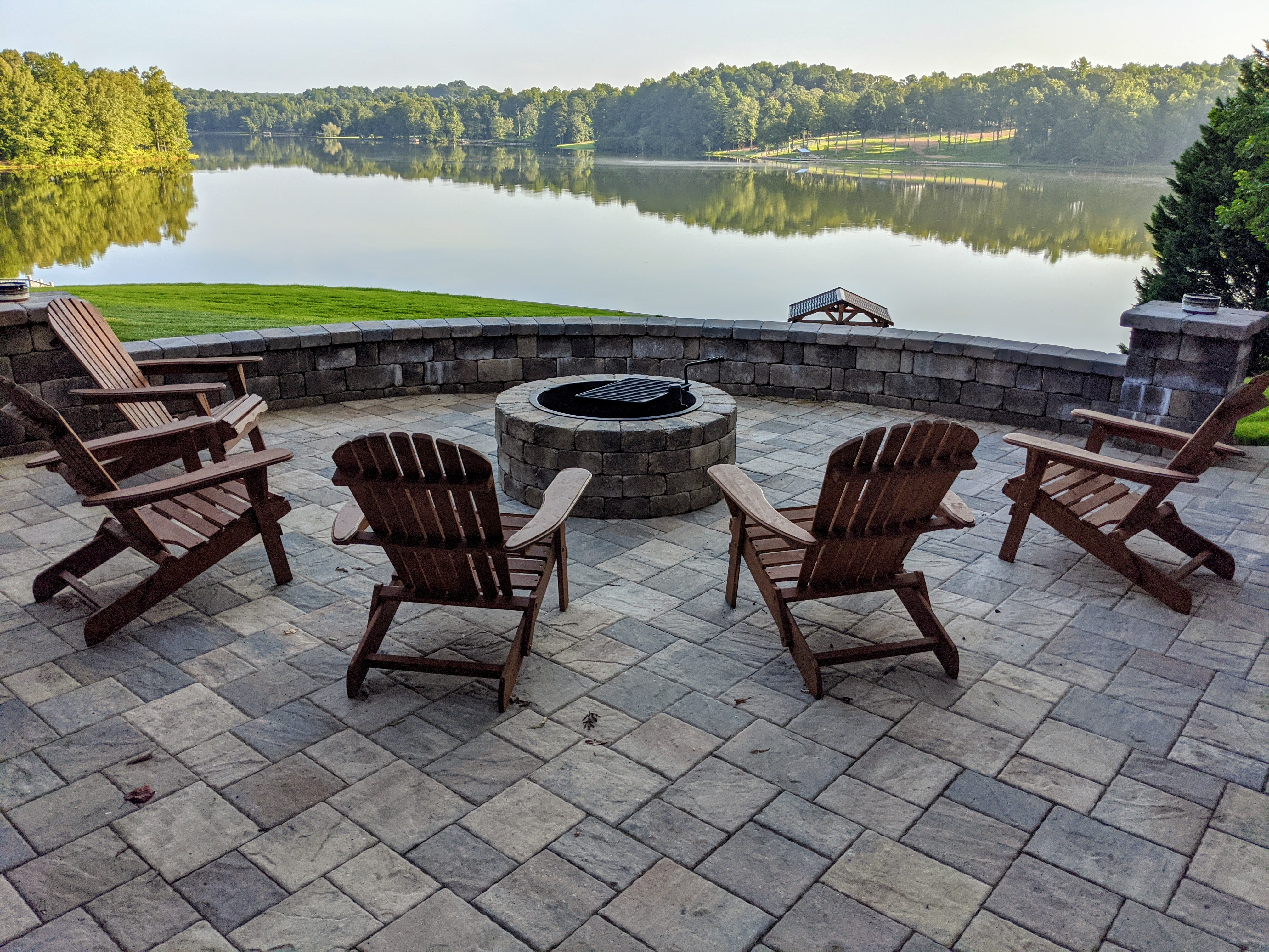 Stone patio with six wooden Adirondack chairs around a fire pit overlooking a calm lake and trees.