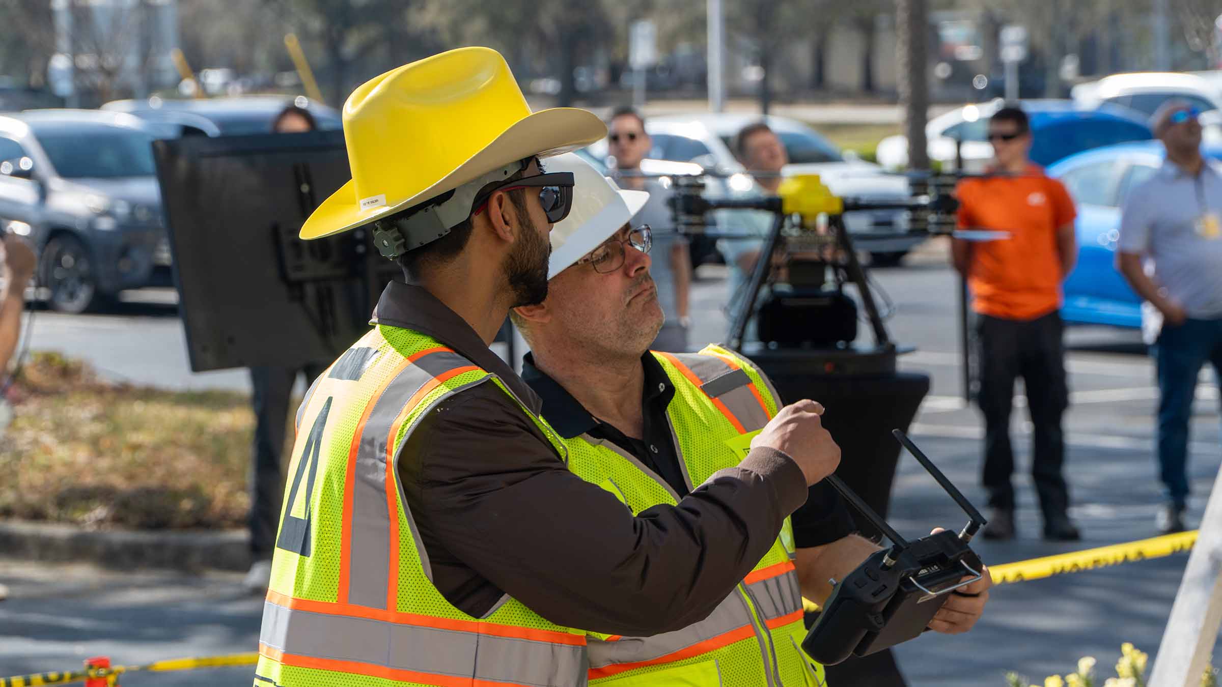 Two construction workers in safety vests and hard hats operating a drone controller outdoors.