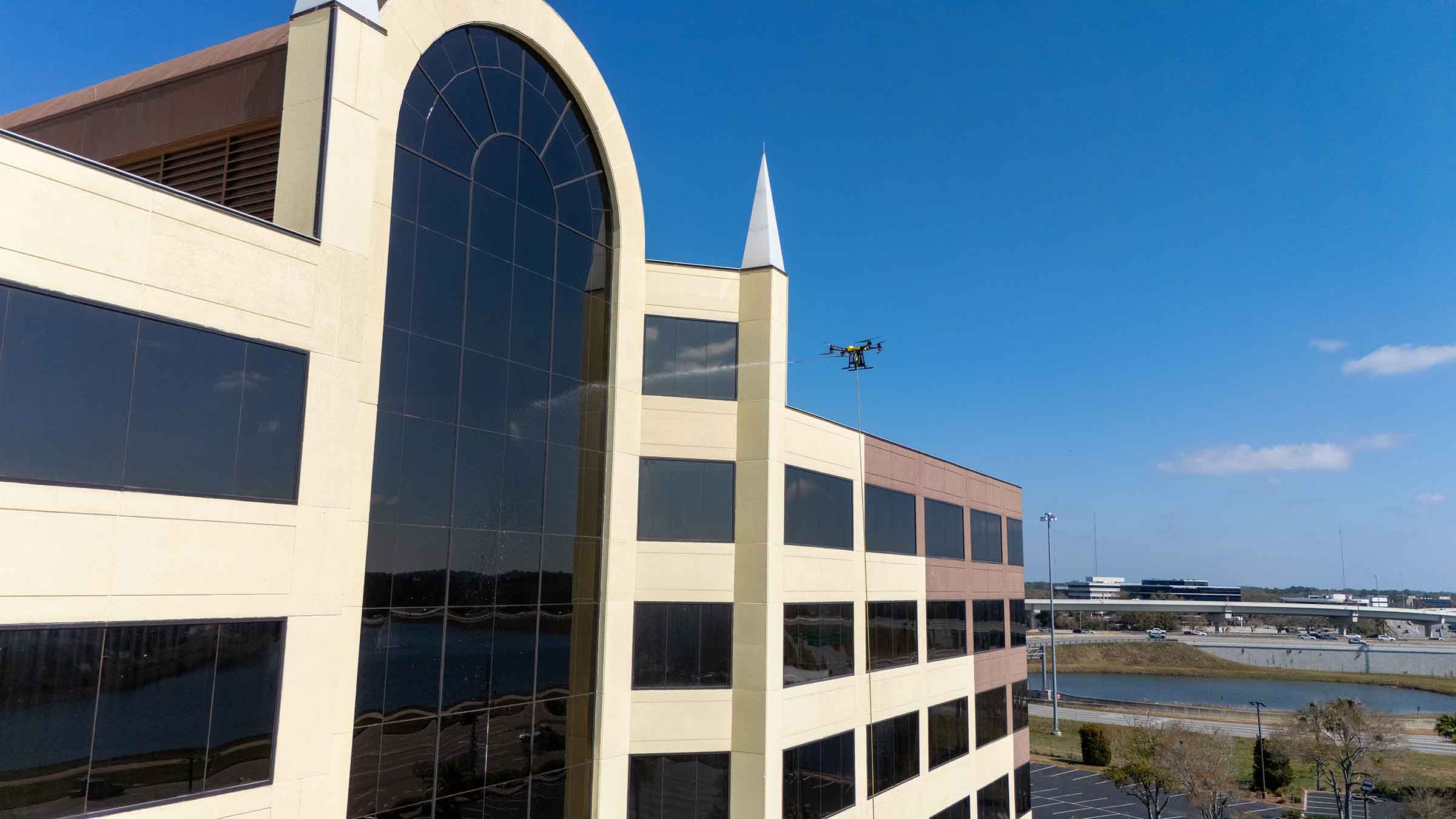 Drone spraying cleaning solution on windows of a multi-story office building under a clear blue sky.