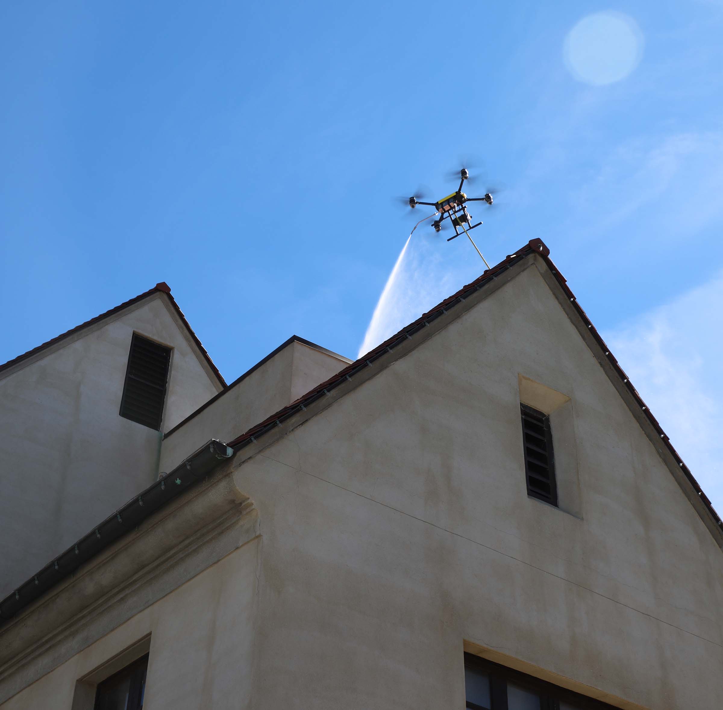 Drone spraying water or cleaning solution on a building roof under a clear blue sky.