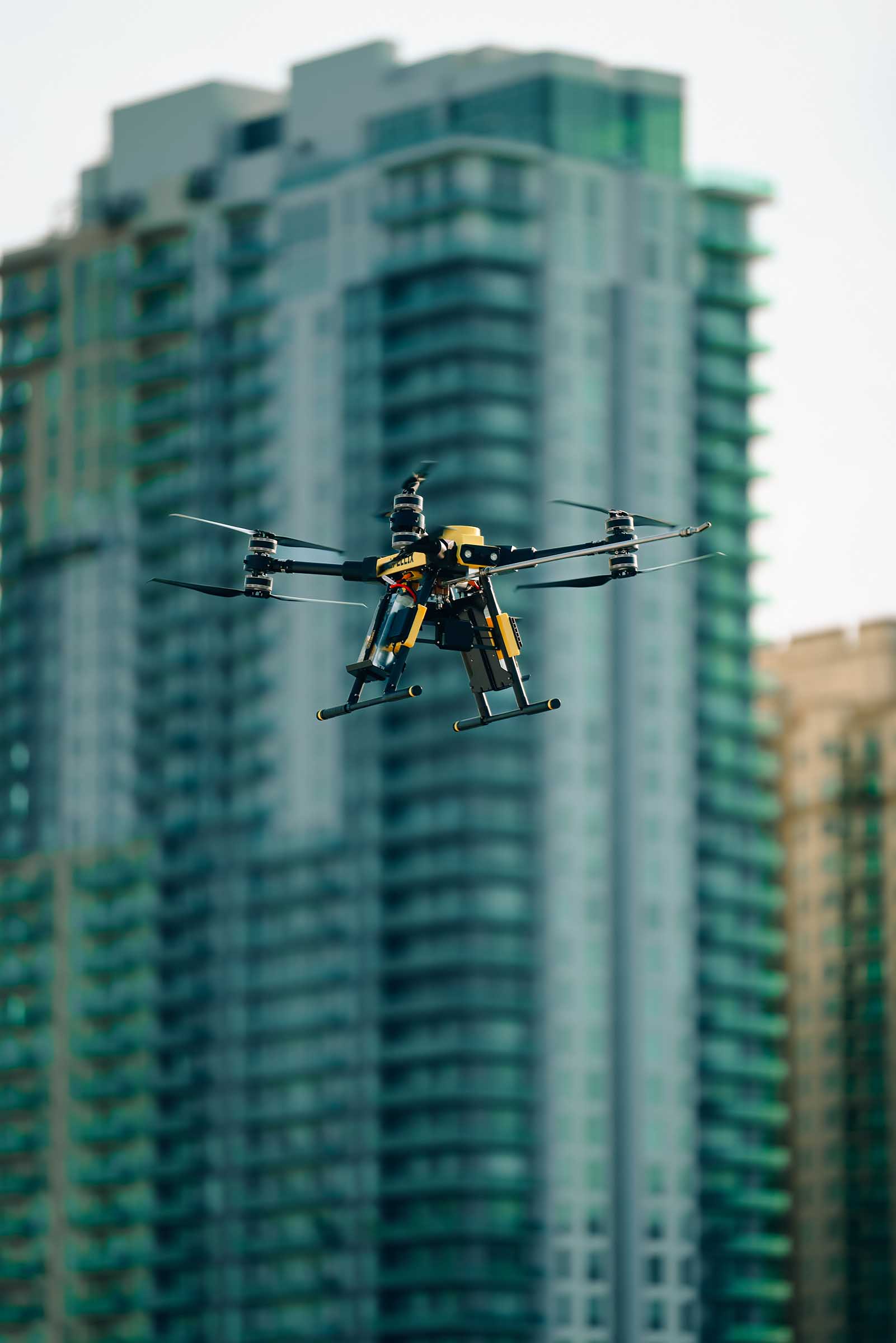 A black and yellow quadcopter drone flying in front of tall urban high-rise buildings.