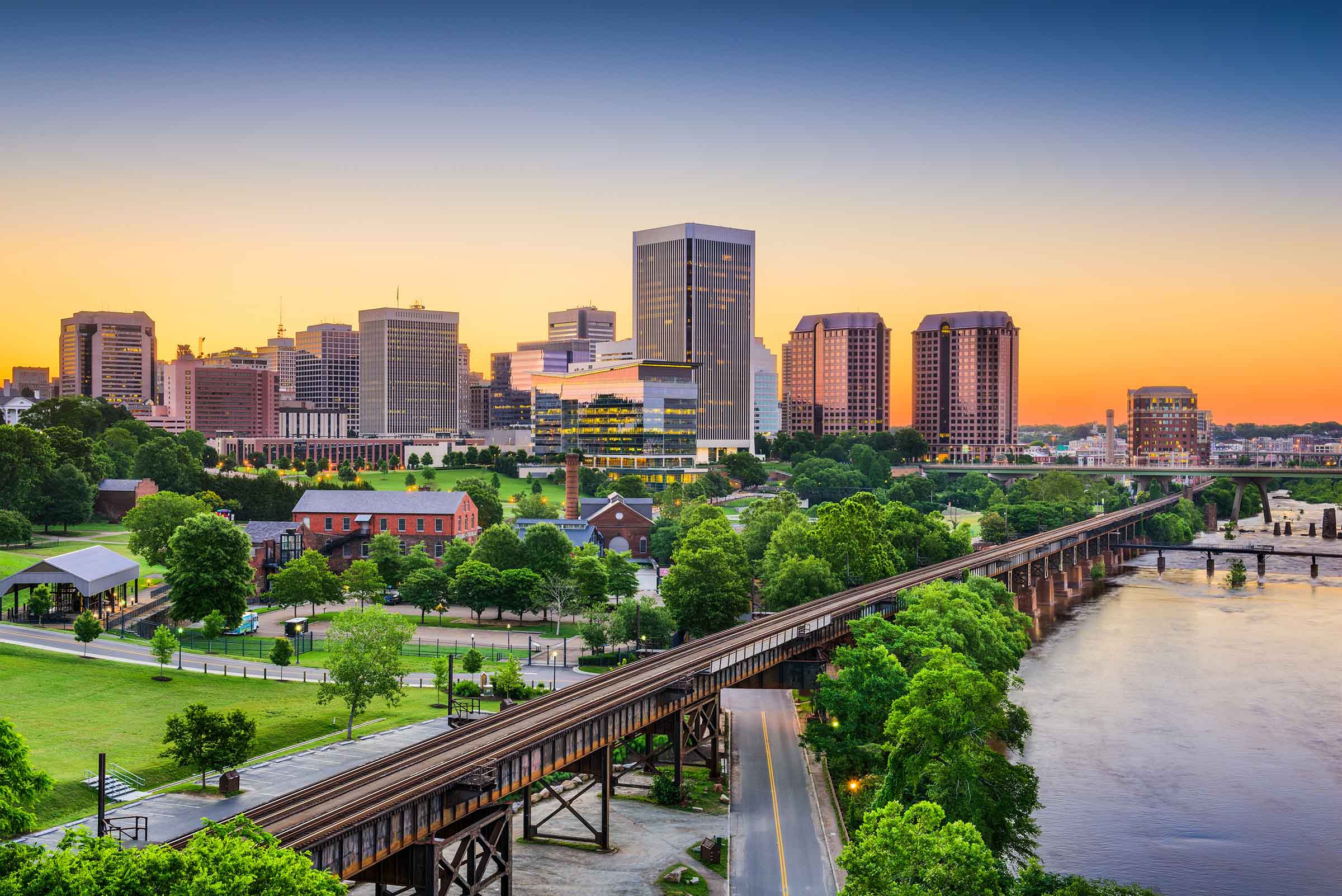 City skyline at sunset with tall buildings, a river, a railway bridge, and lush green trees in the foreground.