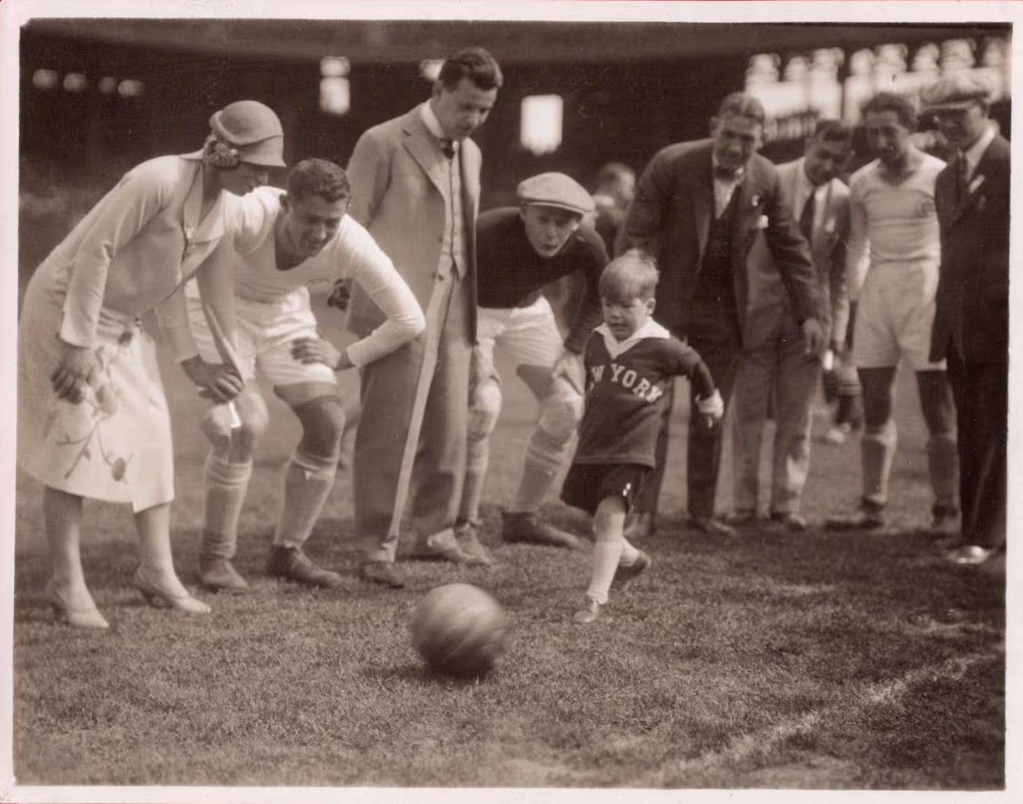 Black-and-white vintage photo of a young boy kicking a soccer ball on a field while a group of adults and players watch closely.