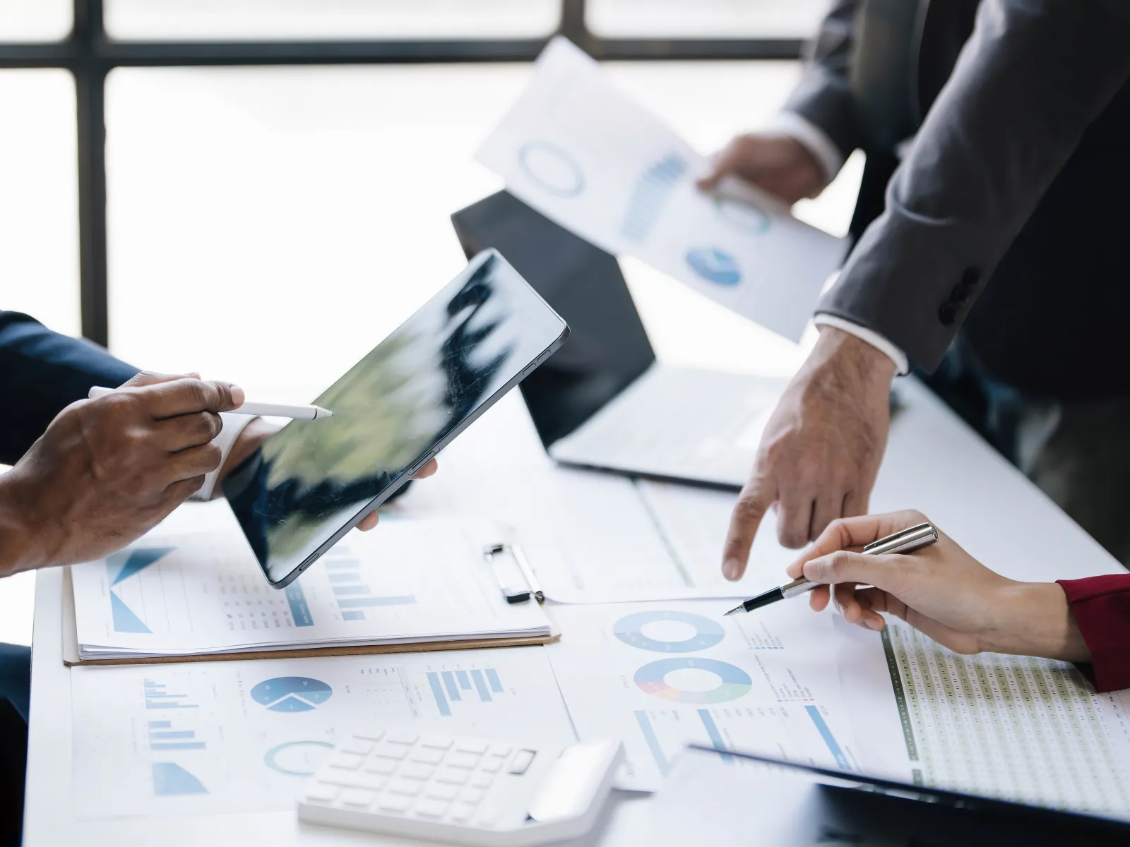 Business professionals reviewing financial charts and reports during a collaborative office meeting, with one person using a tablet and others pointing at printed analytics documents on a conference table.