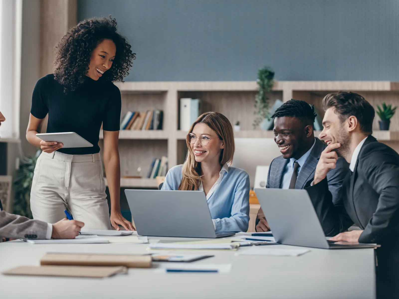 Diverse team of business professionals collaborating in a modern office meeting, smiling and discussing ideas around laptops and documents at a conference table.