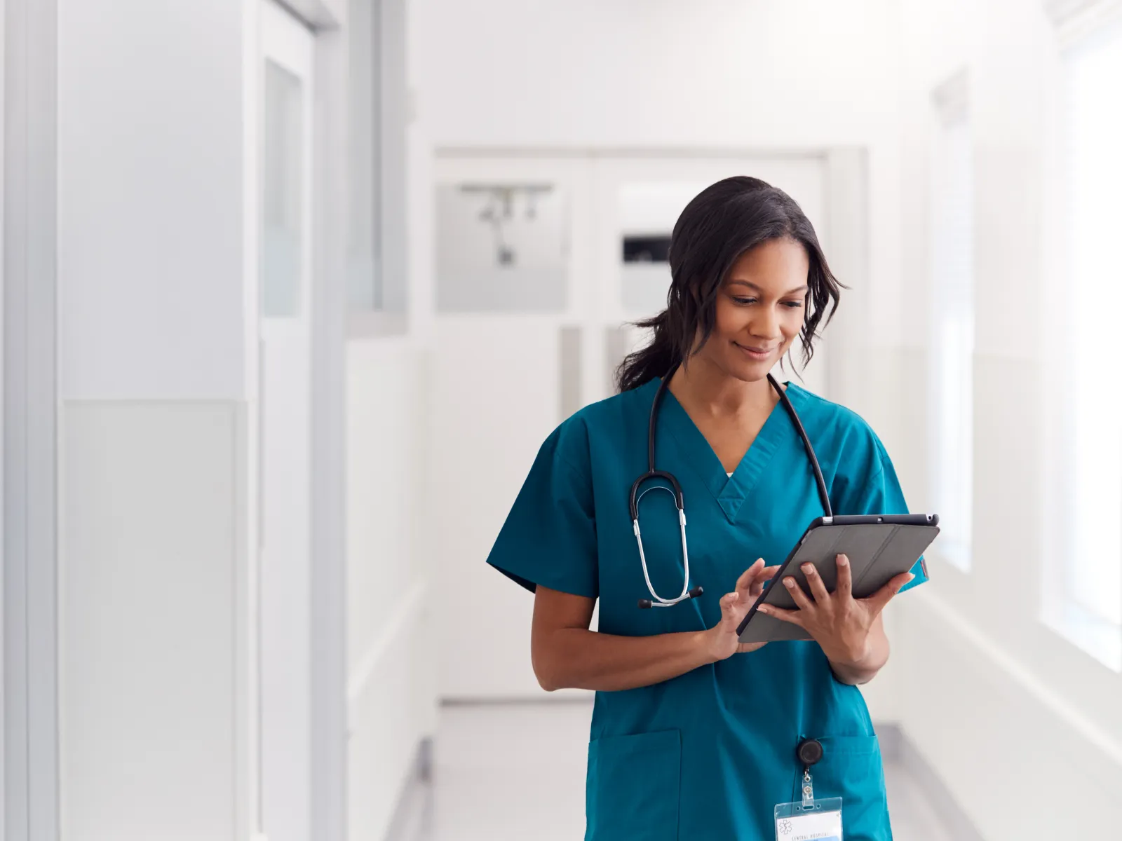 Female nurse in scrubs using tablet in hospital corridor with stethoscope