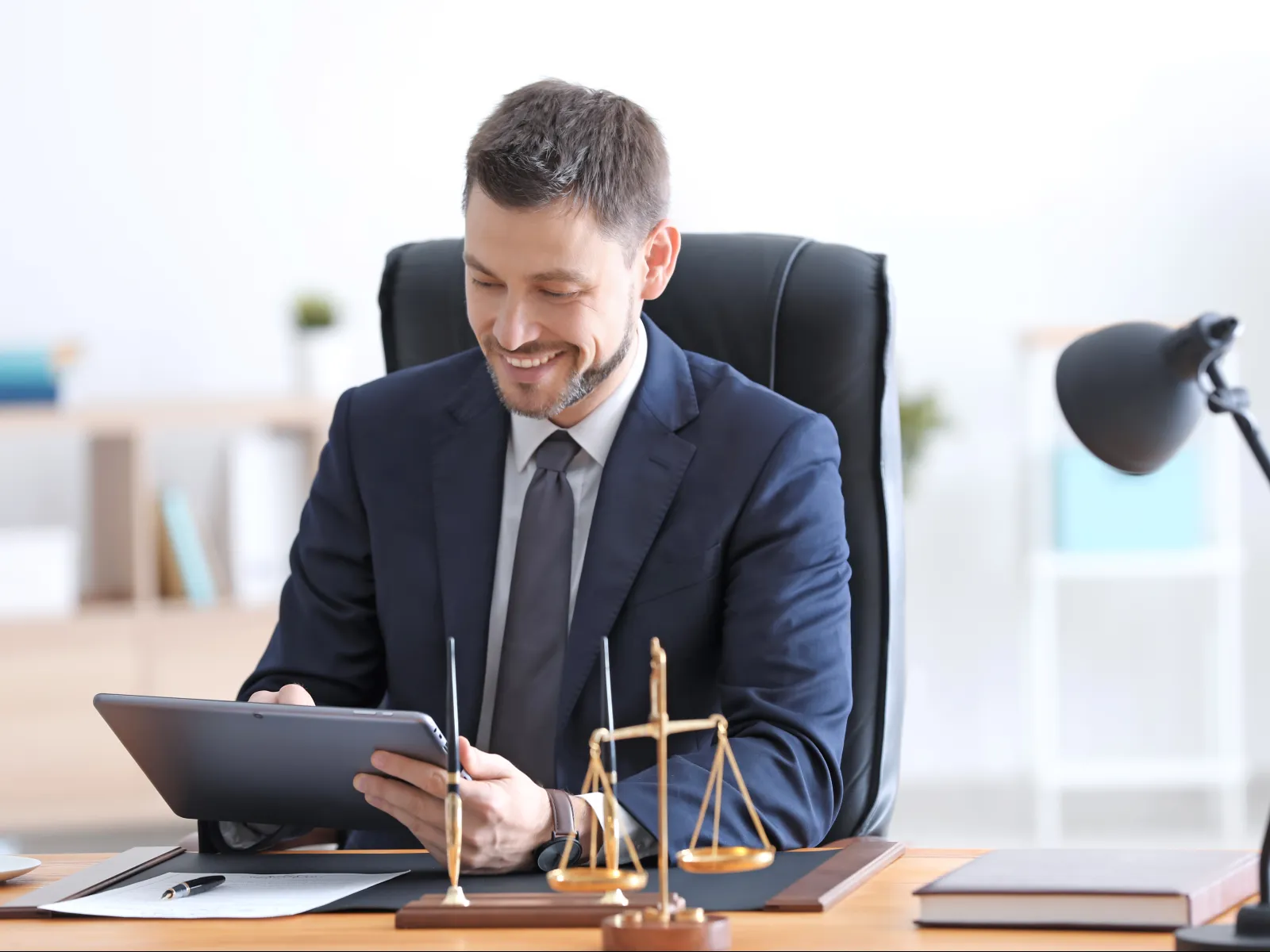 Smiling lawyer working on tablet at desk with scales of justice in modern law office