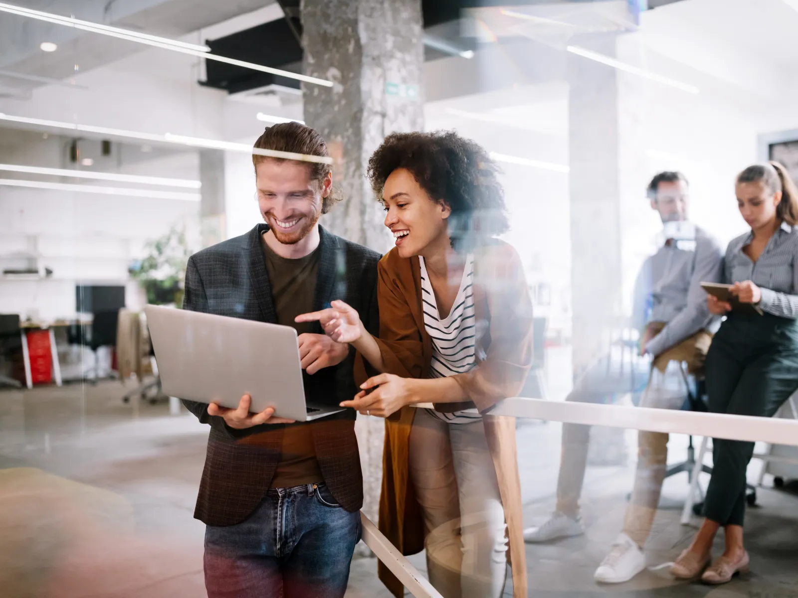 Two colleagues stand together in a modern office, looking at a laptop and smiling, while others work in the background.