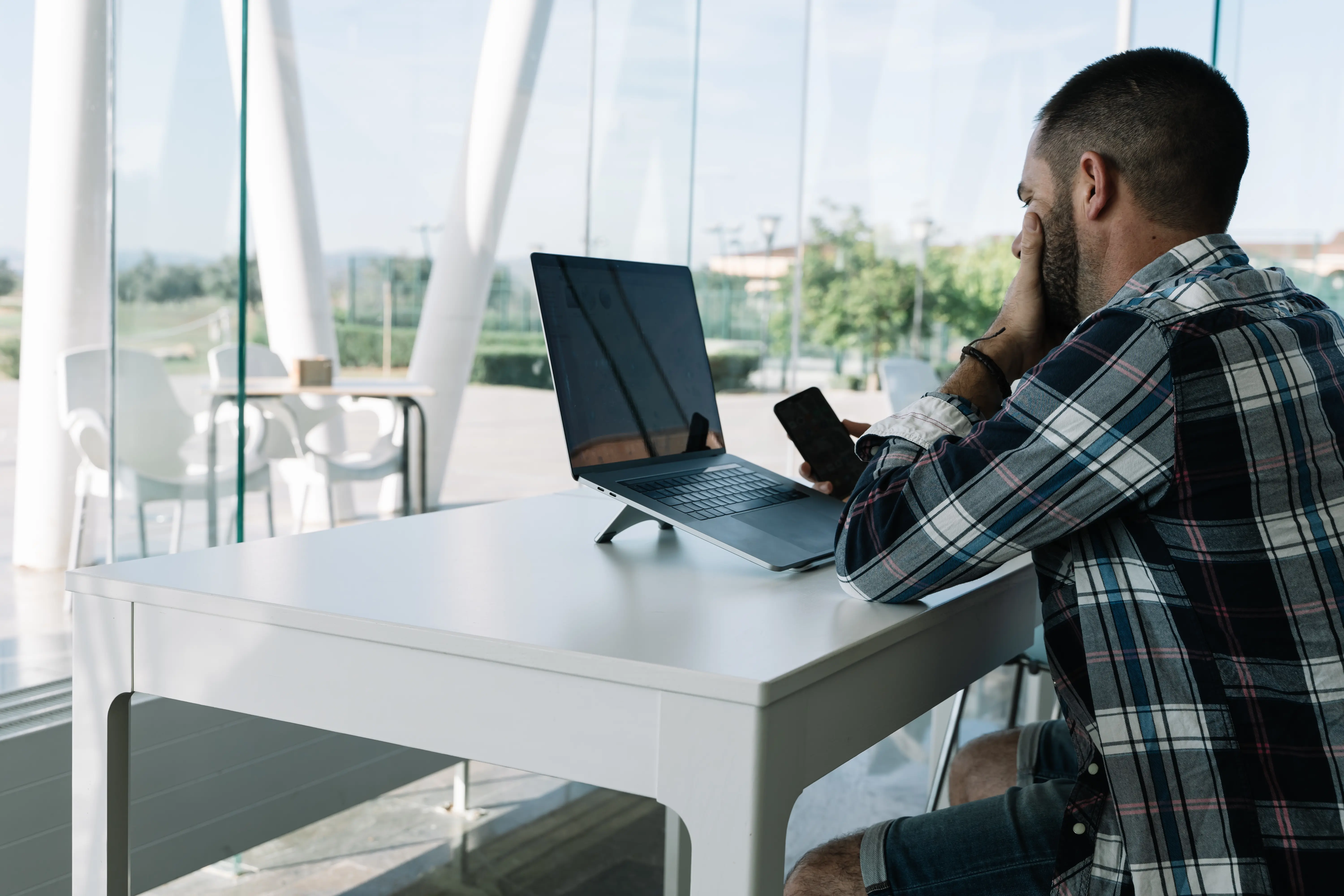 Man holding smartphone while working on laptop at a modern bright workspace with glass walls