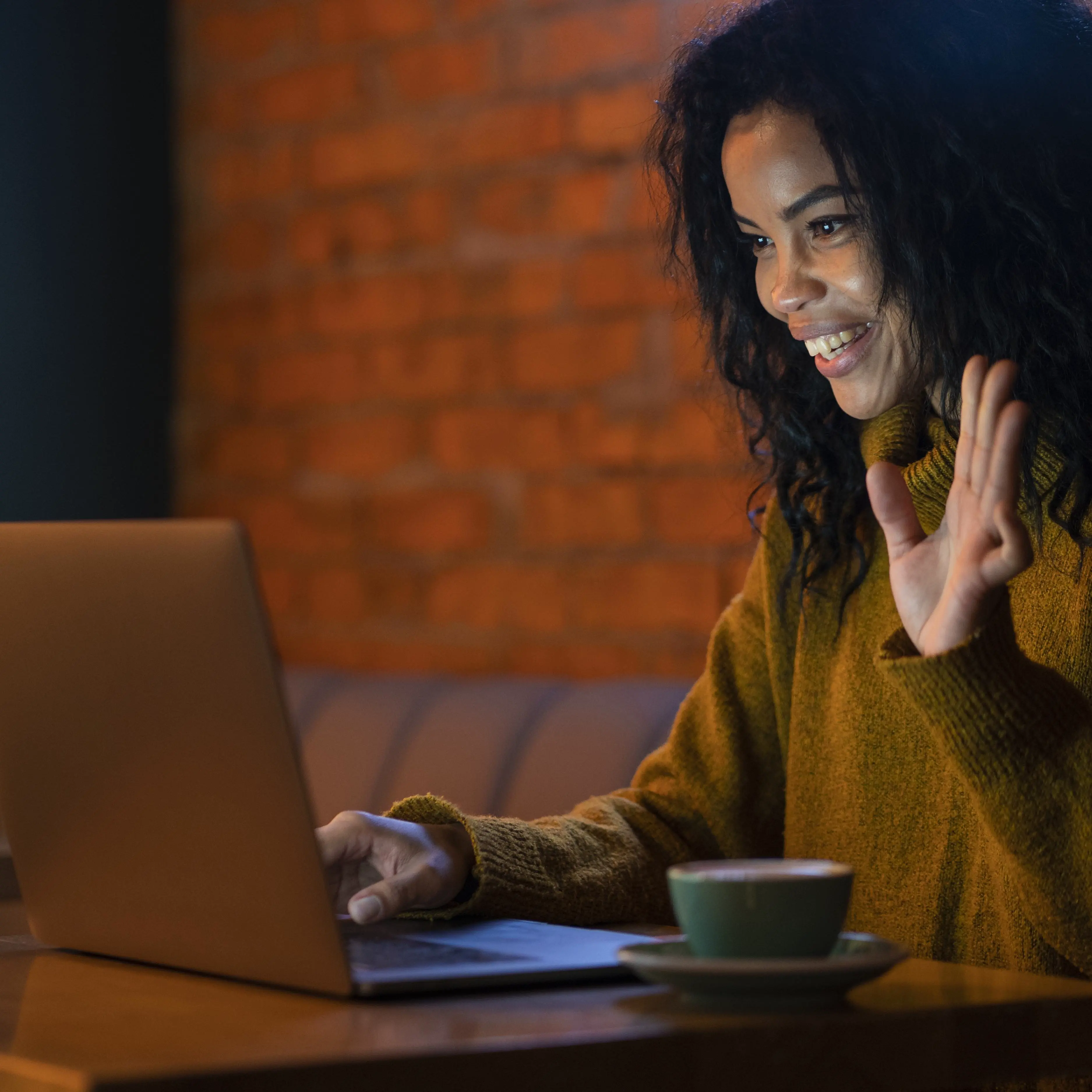 Smiling woman waving during a video call on laptop at a cozy coffee shop with brick wall background