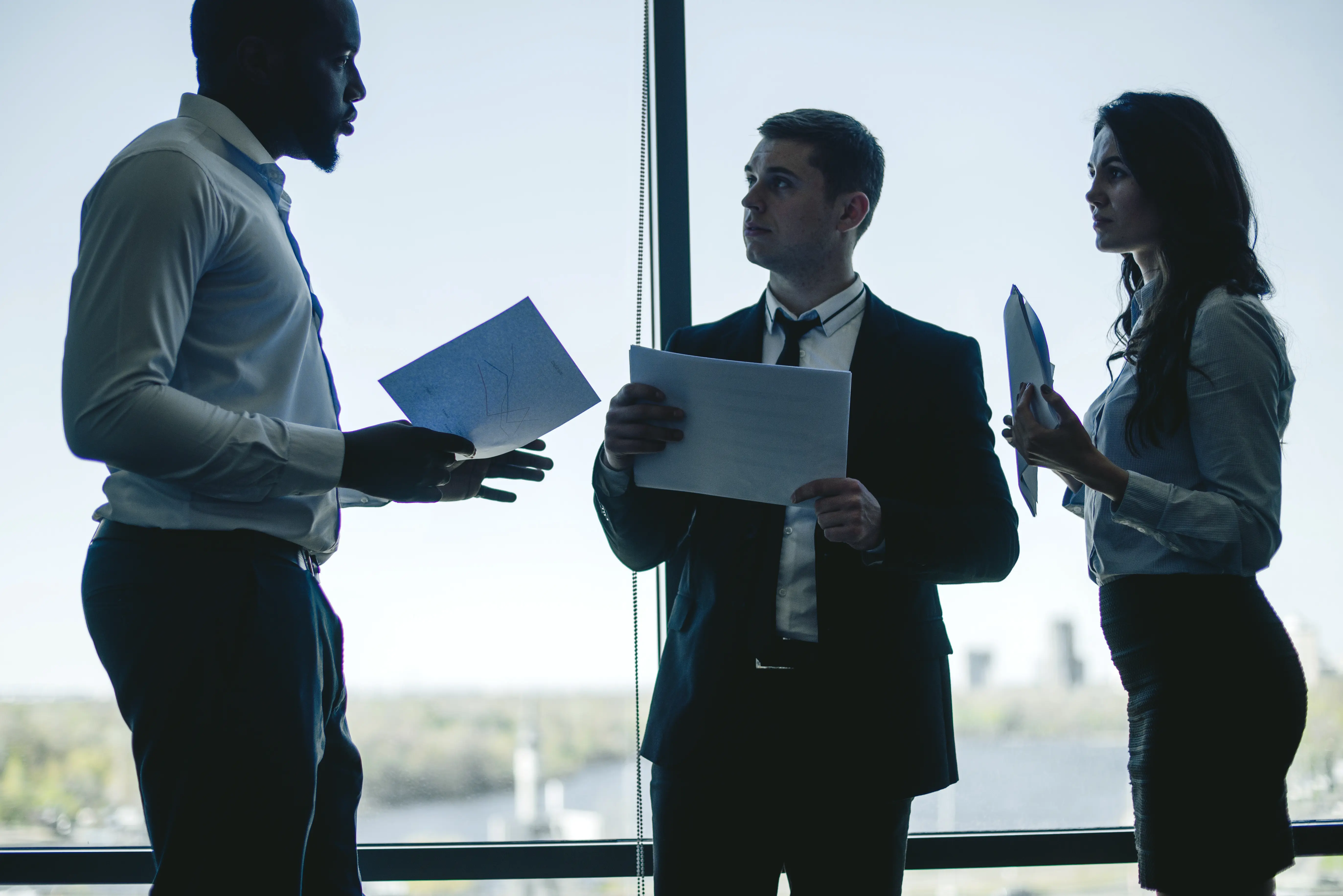 Three business professionals discussing documents in silhouette against large office windows