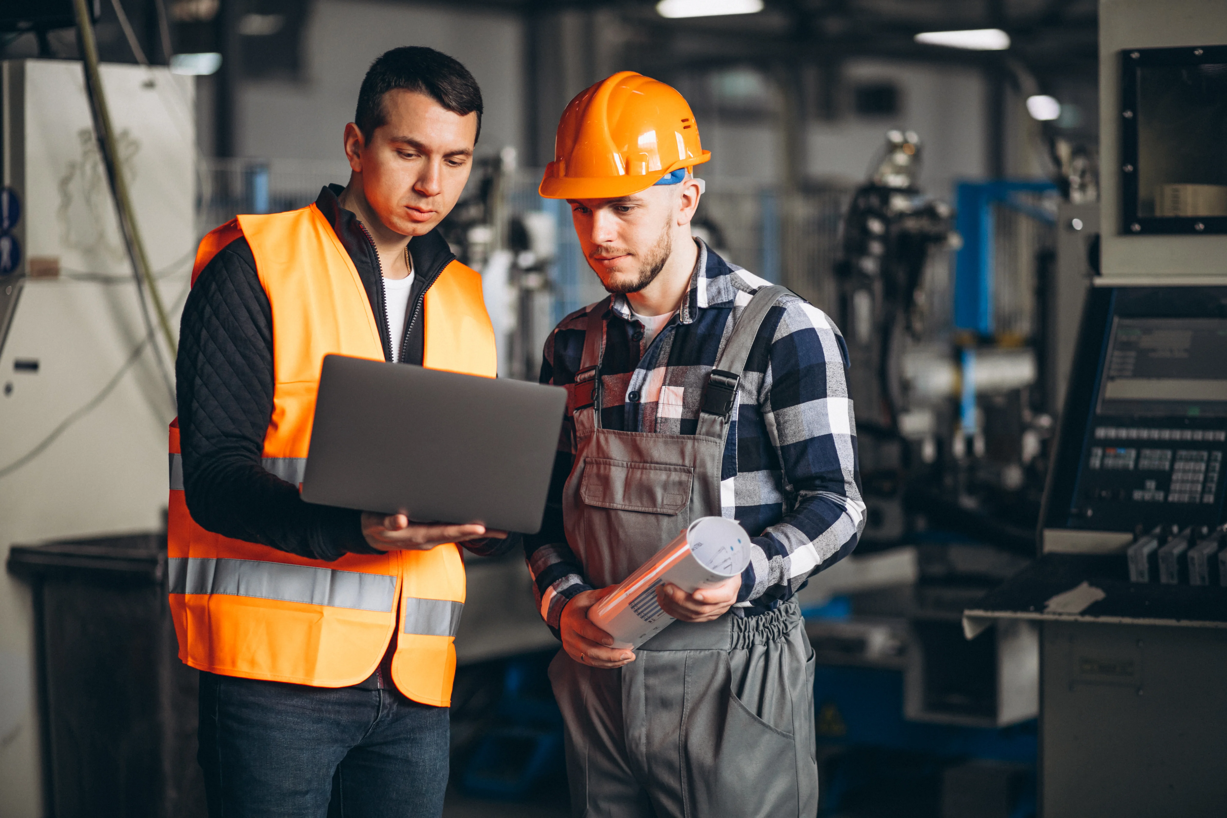 Two factory engineers reviewing data on a laptop on the production floor, one in a safety vest and one in a hard hat