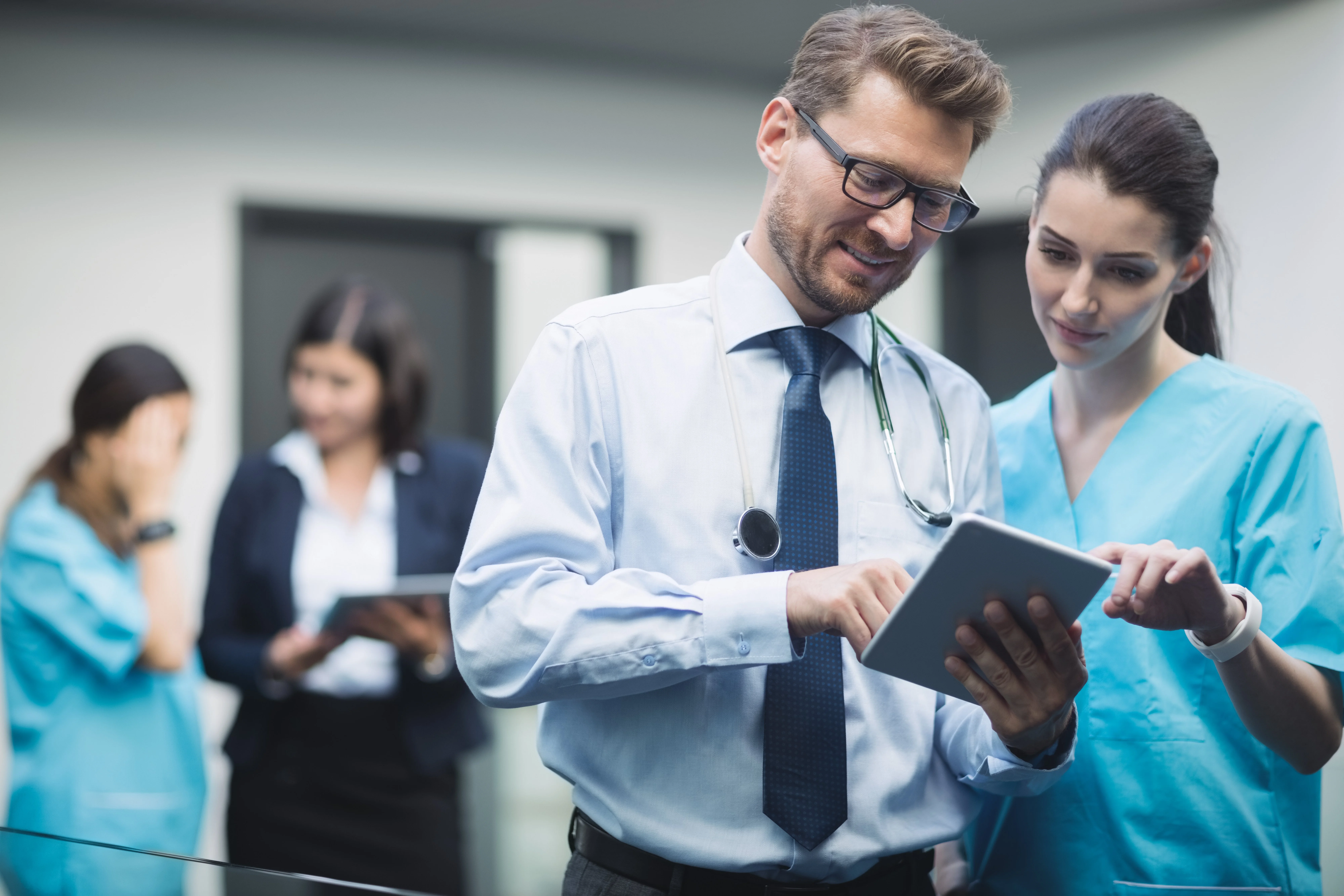 Doctor and nurse reviewing patient information on a digital tablet in a hospital corridor, with colleagues in the background