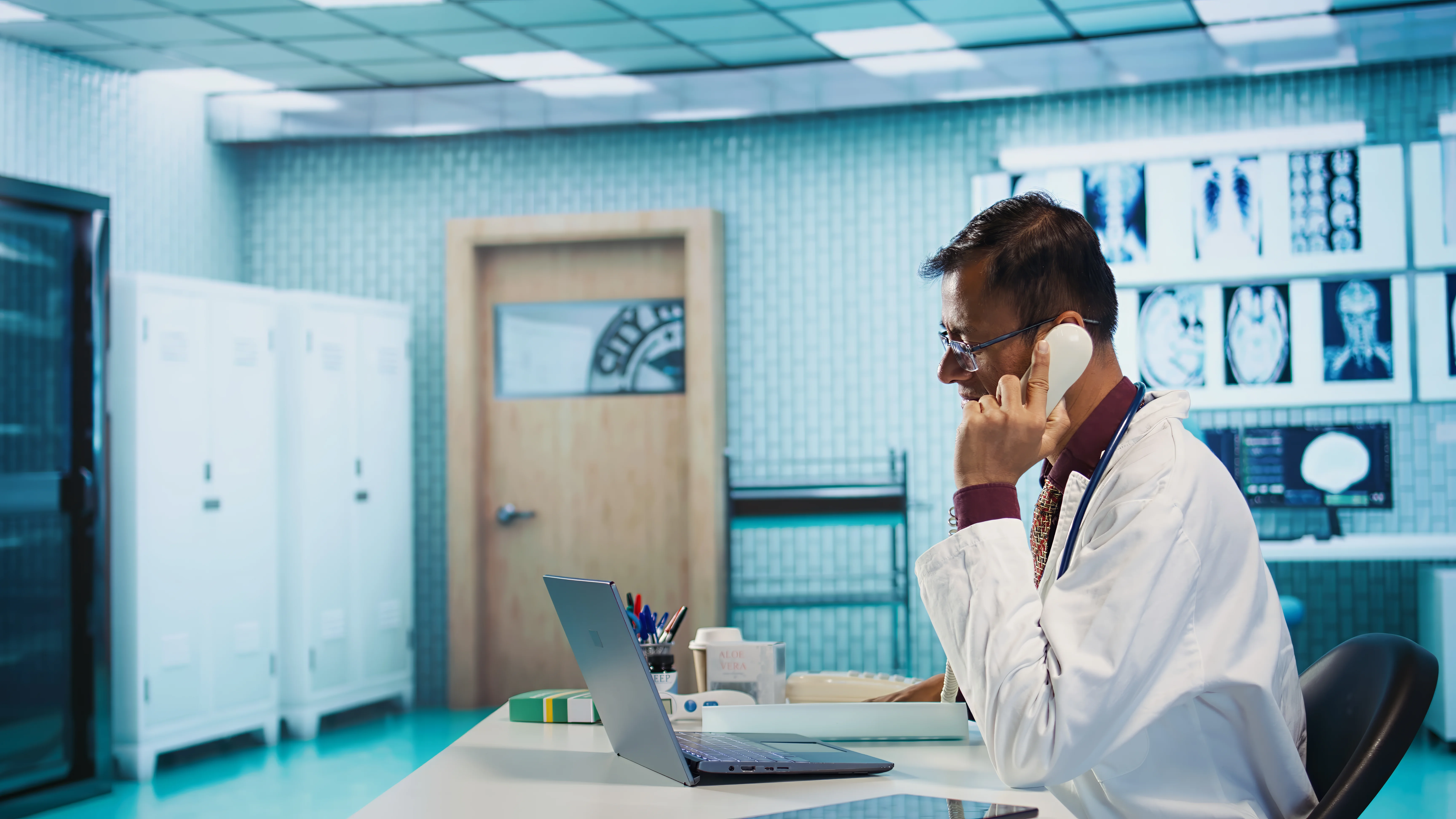 Doctor on the phone using a laptop in a medical office, with X-ray and MRI scan images displayed on the wall behind him