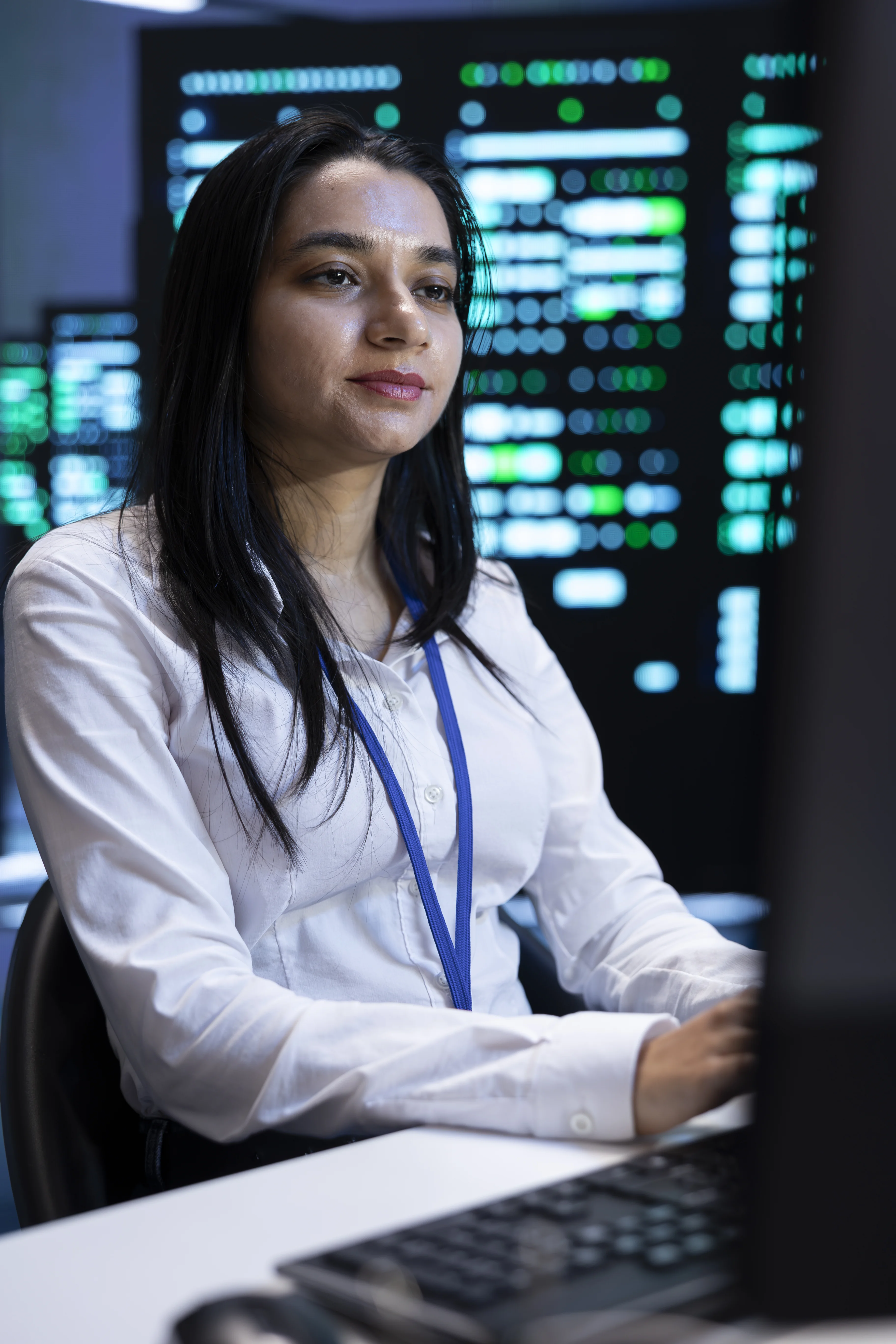 Data center engineer working at a computer in front of large monitoring screens showing green and blue system status displays