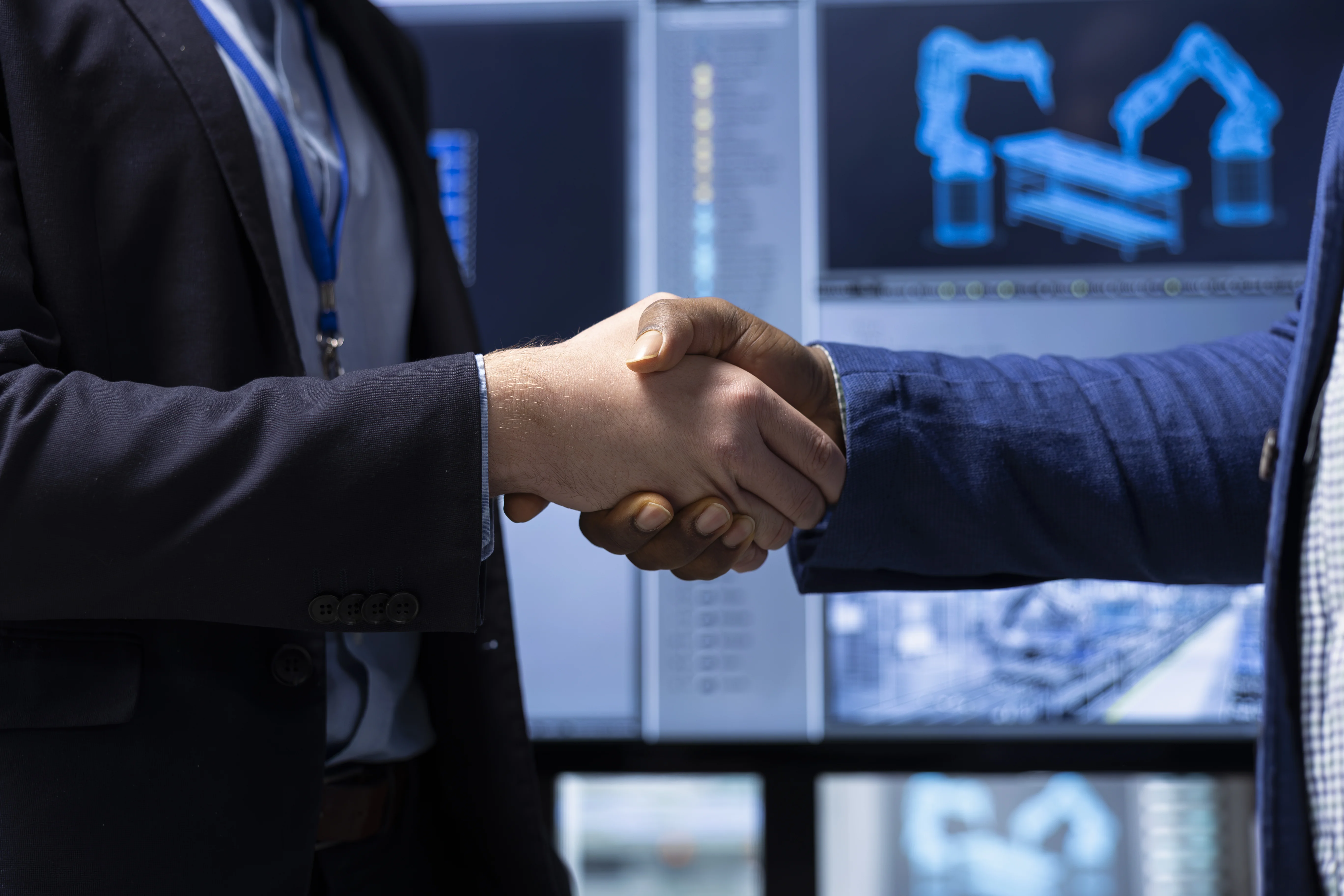 Close-up of two business professionals shaking hands in front of screens showing robotic automation diagrams in a factory