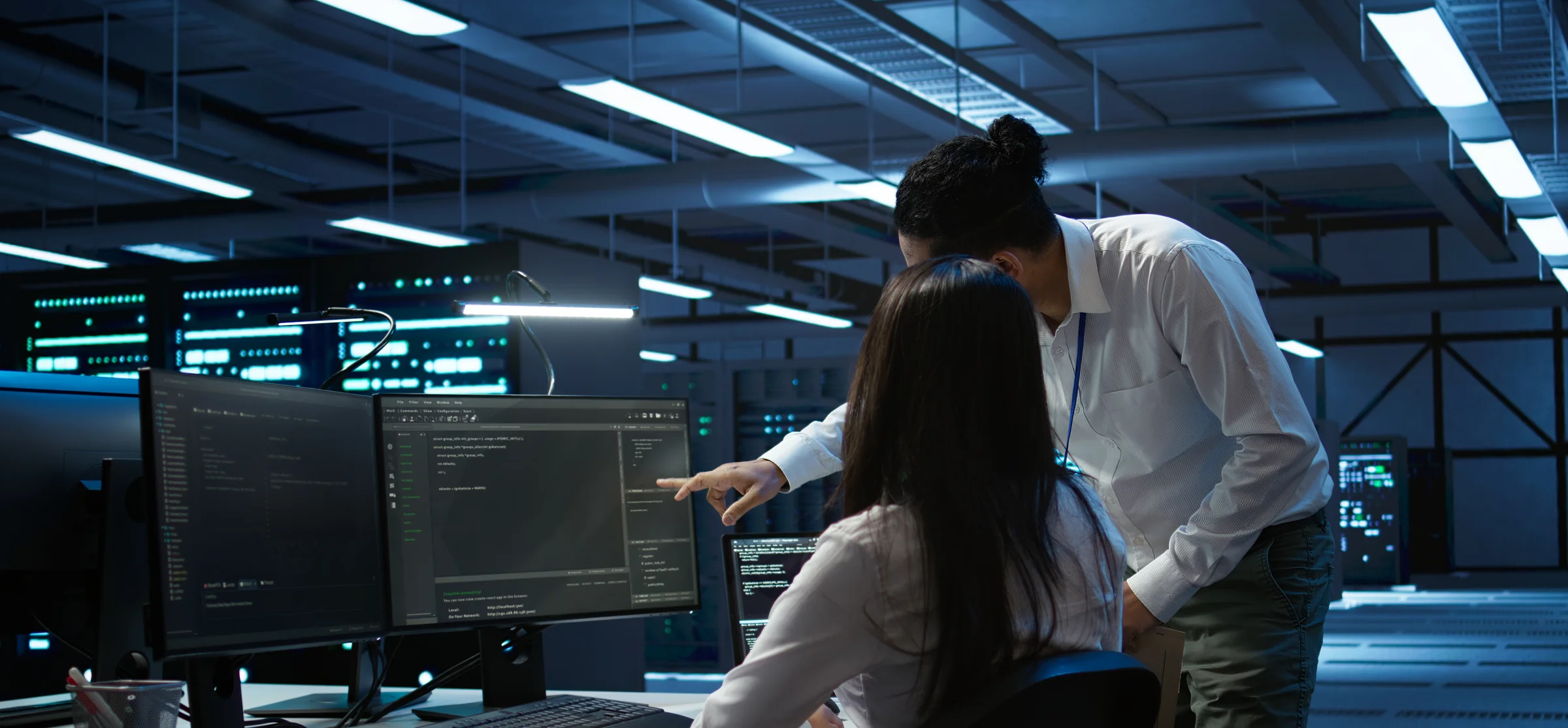 Two IT professionals reviewing code on a monitor together in a data center, with rows of servers glowing in the background