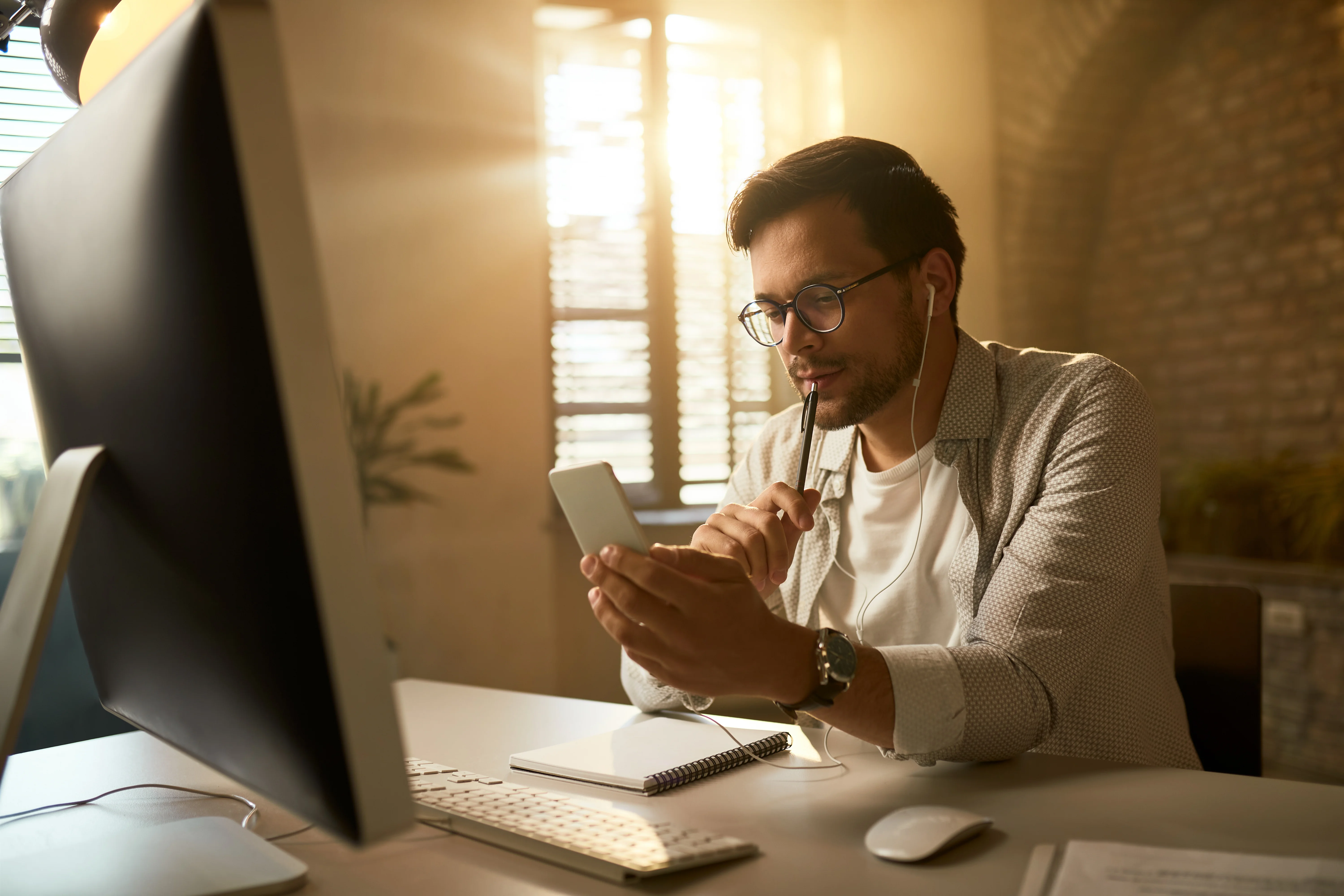 Man in glasses with earphones using a smartphone at his desk beside a computer in a warmly lit home office