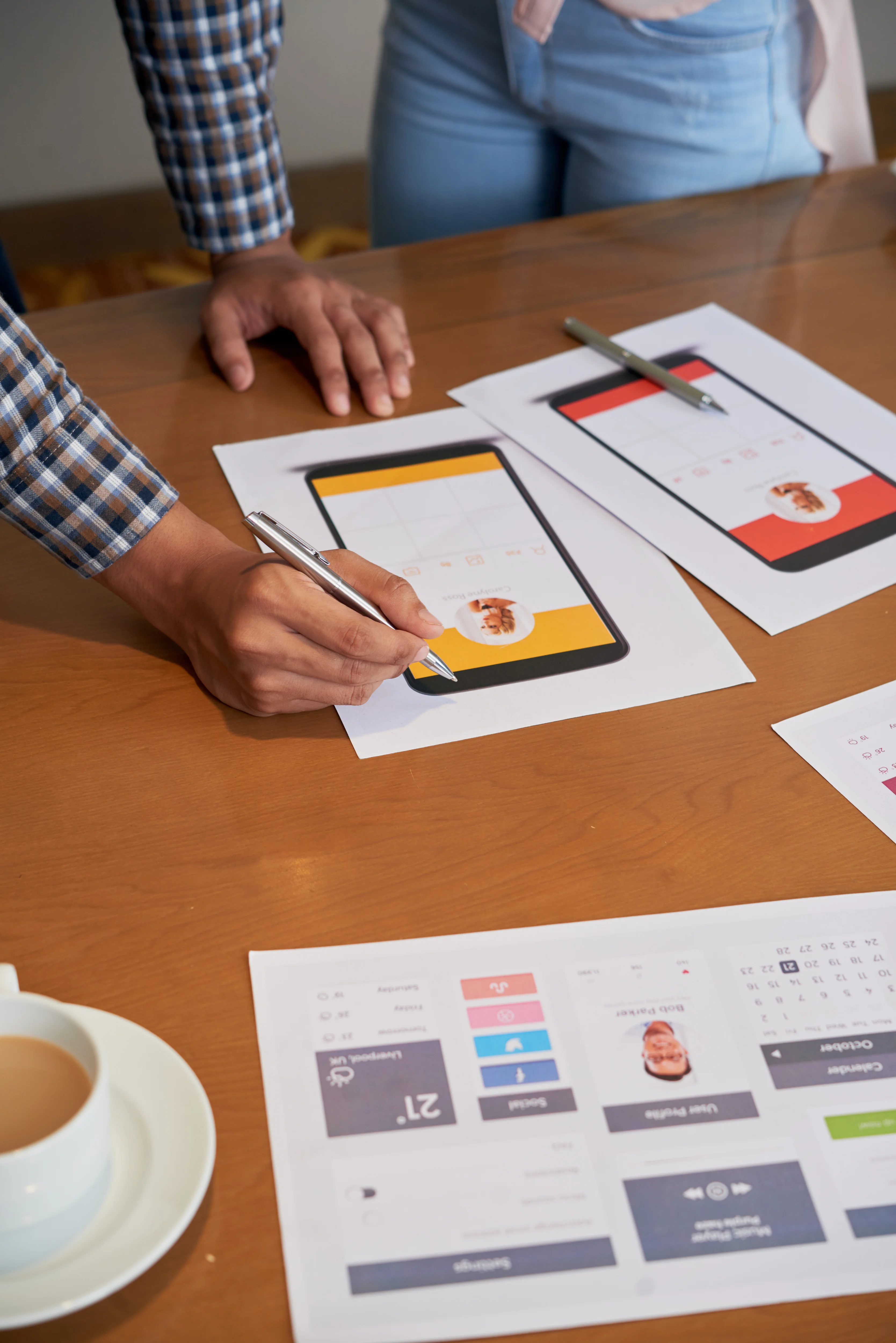 Two designers reviewing printed mobile app UI mockups and wireframes on a table during a design meeting
