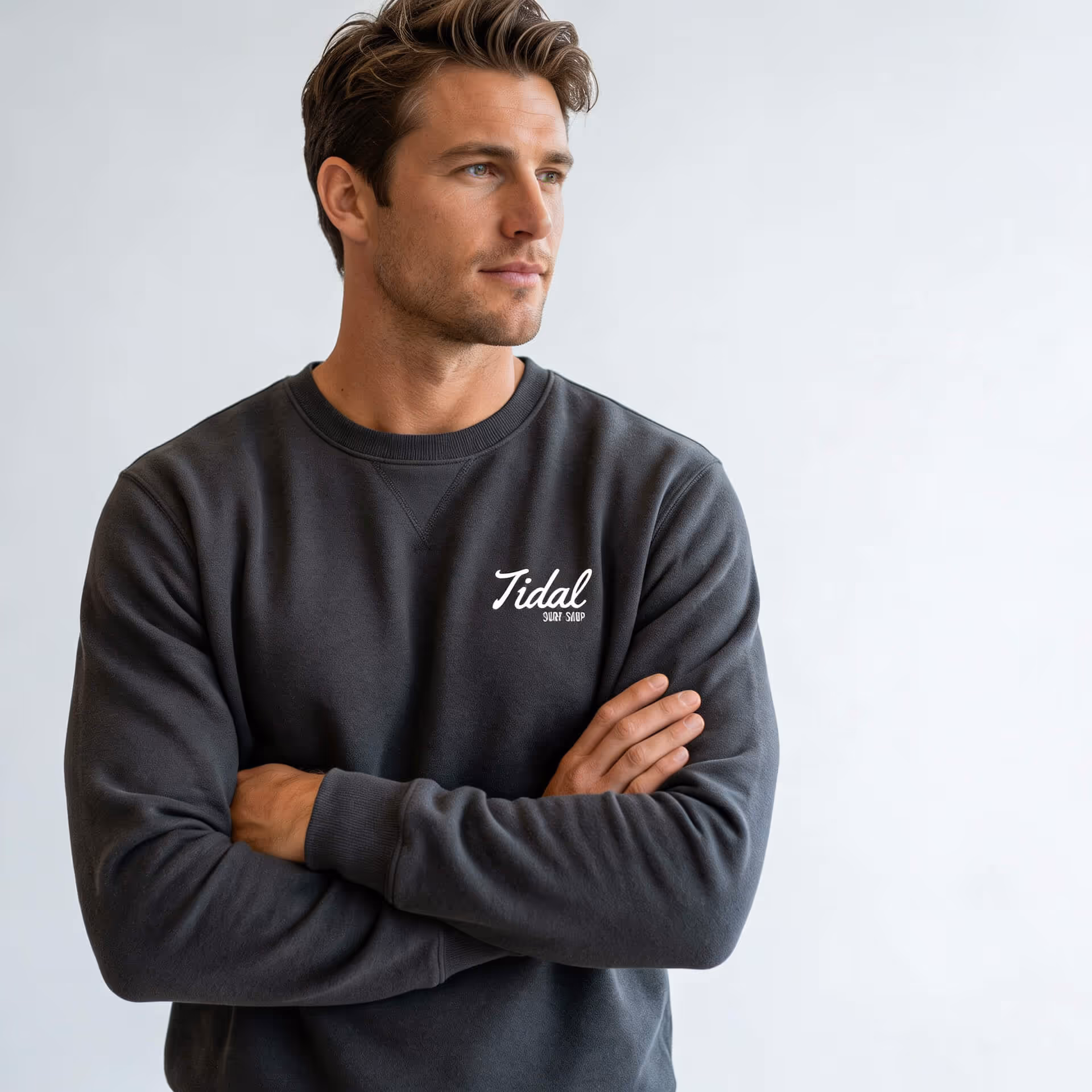 Man wearing dark gray sweatshirt with Tidal Surf Shop logo, arms crossed, looking to the side.