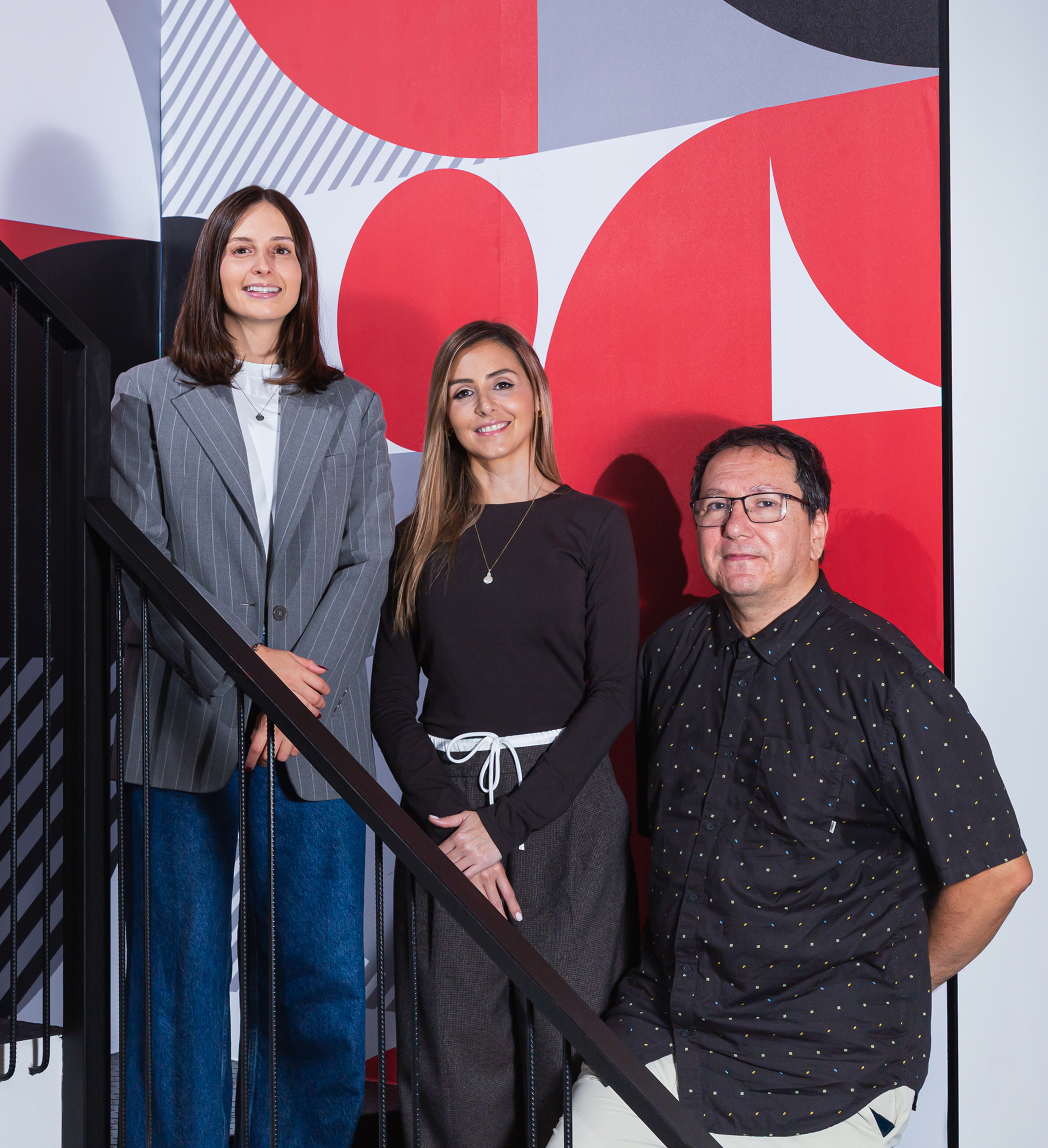 Tres personas sonrientes de pie junto a una barandilla negra frente a una pared con arte geométrico rojo y gris.