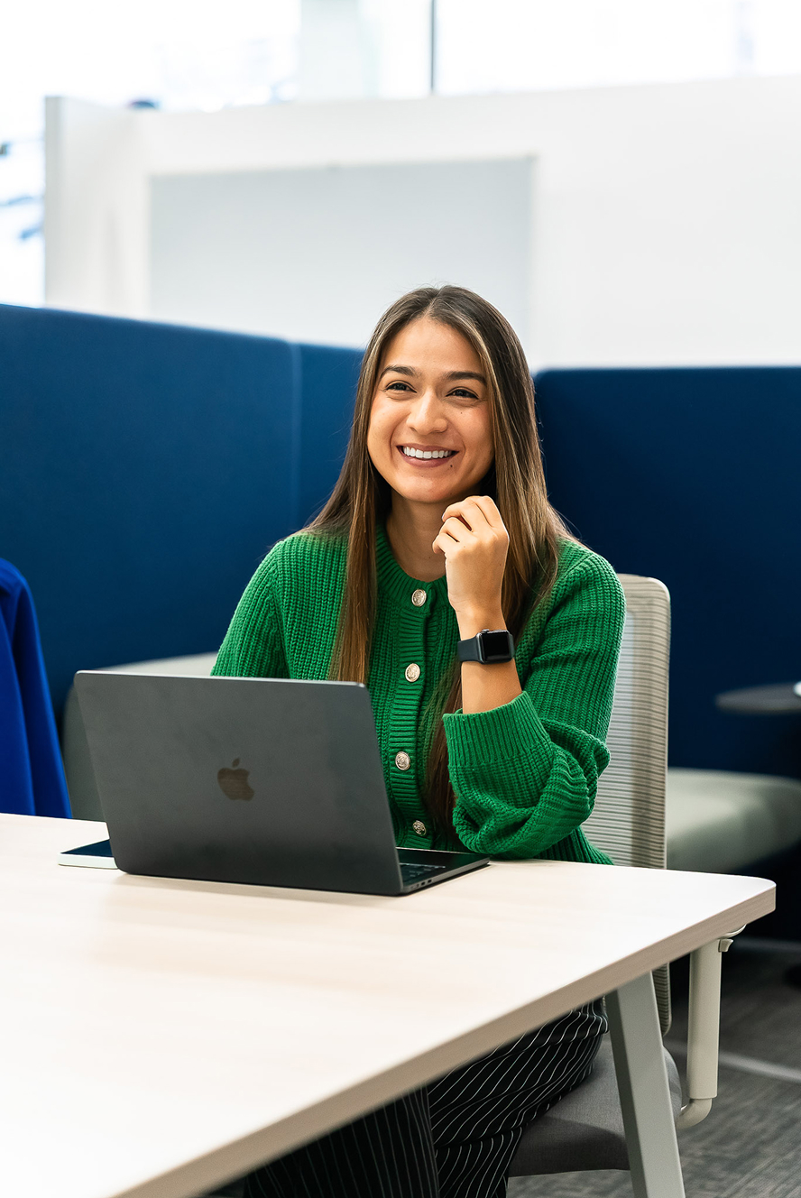 Mujer sonriente con suéter verde trabajando en una computadora portátil en una oficina moderna.