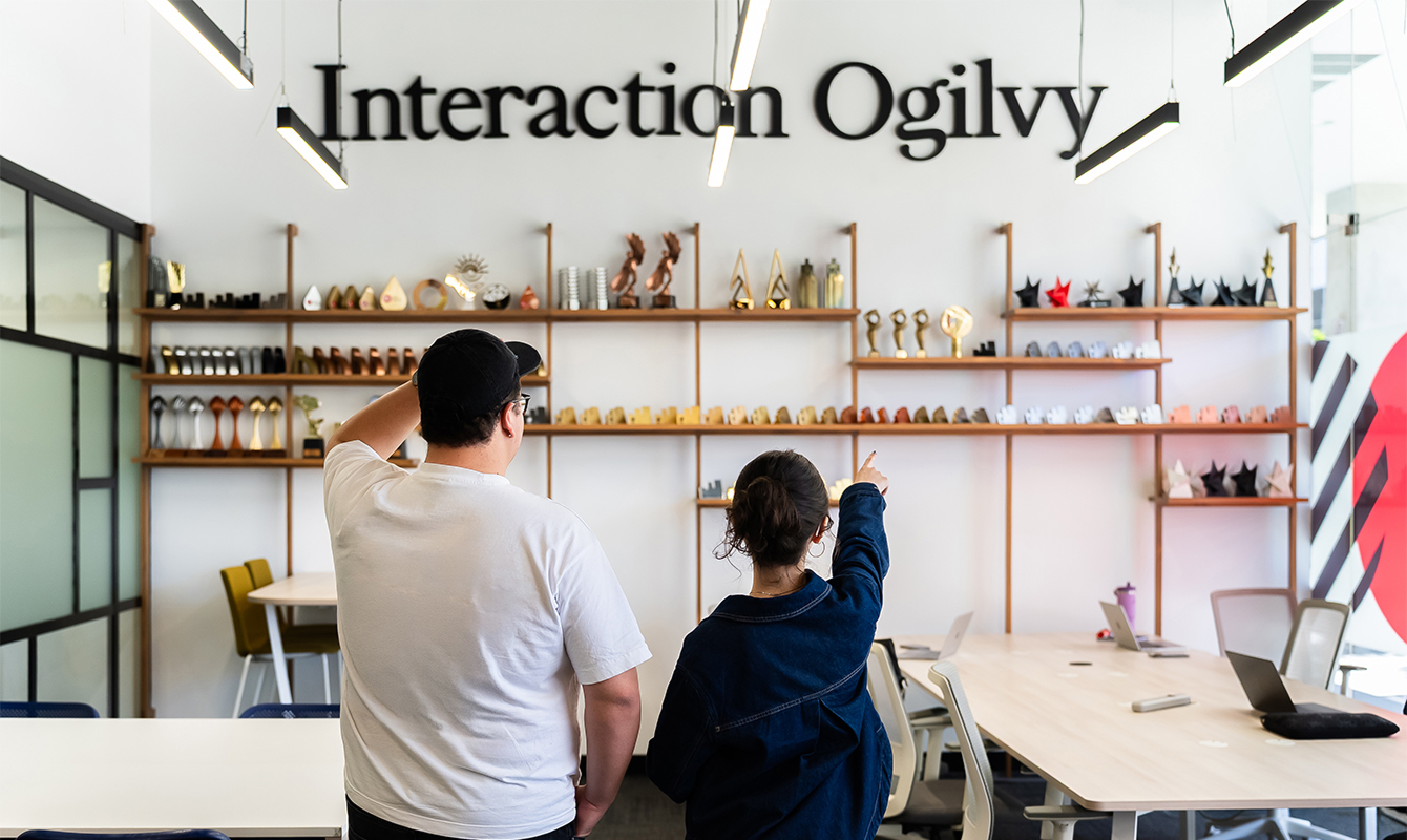 Dos personas de espaldas observan una pared con trofeos y la inscripción Interaction Ogilvy en una oficina moderna.