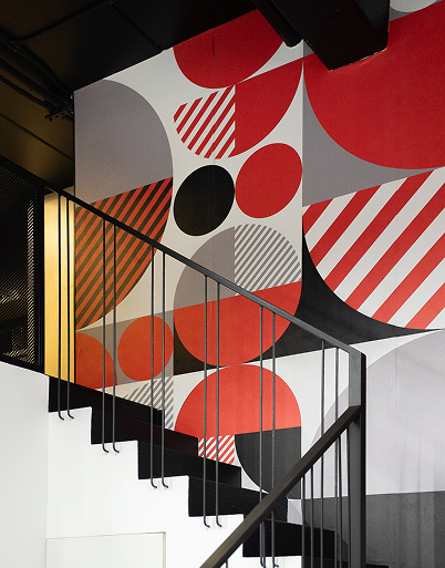 Escalera negra con barandilla metálica frente a una pared con arte geométrico en tonos rojo, negro y gris.