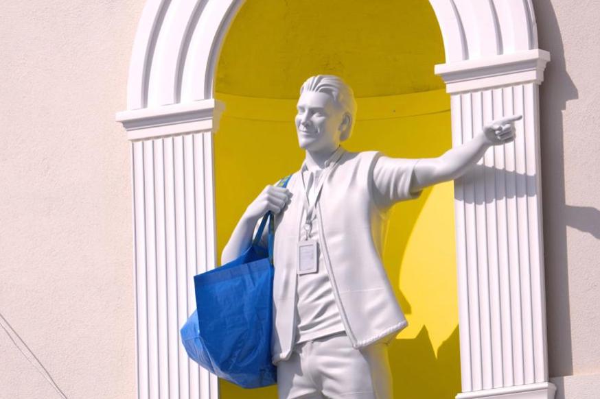Estatua blanca de hombre sonriendo con bolso azul y gafete señalando hacia la derecha frente a fondo amarillo.