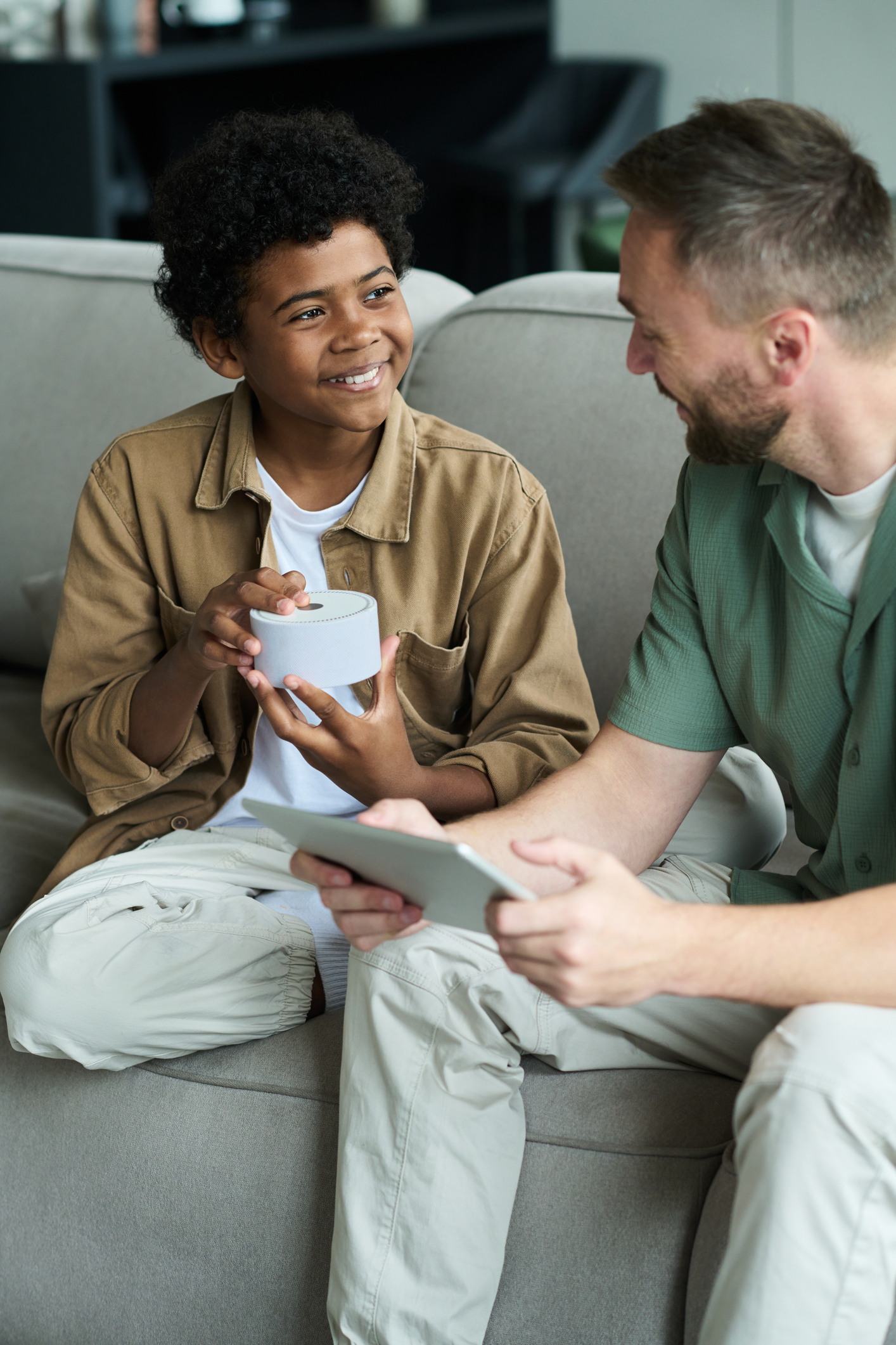 Young girl in a red shirt writing on paper while sitting among other children in a classroom with sunlight coming through window blinds.