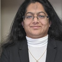Smiling woman with long black hair, glasses, white turtleneck, black blazer, and gold necklace.